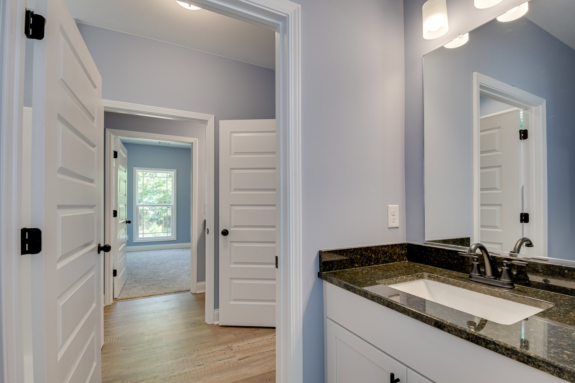 Bathroom with white countertop and undermount sink, chrome faucet, large framed mirror above, white cabinetry, tile backsplash, window showing trees outside, white door with black