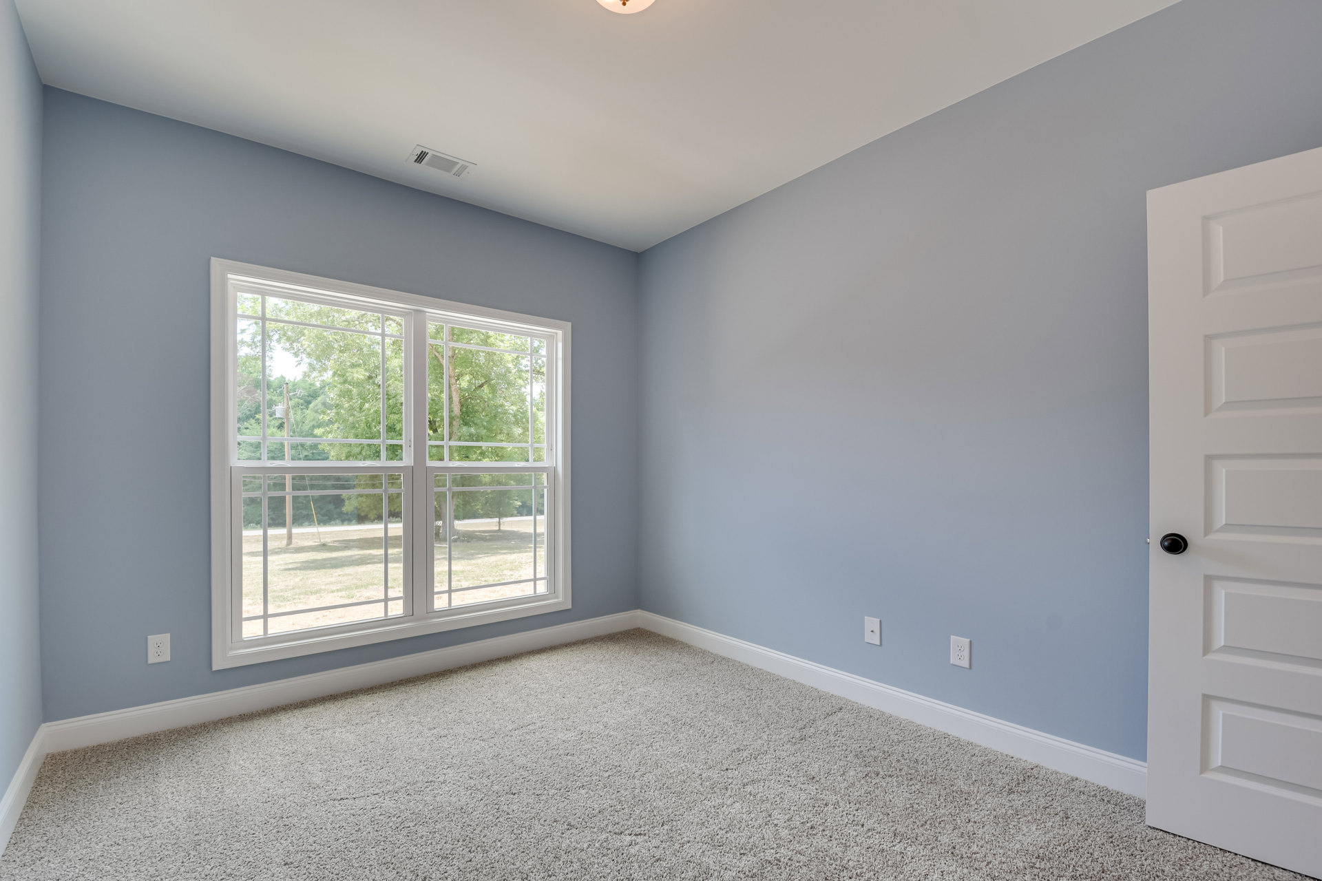 Carpeted room with white walls, large window overlooking trees and grass, white door with black knob, white ceiling featuring recessed light
