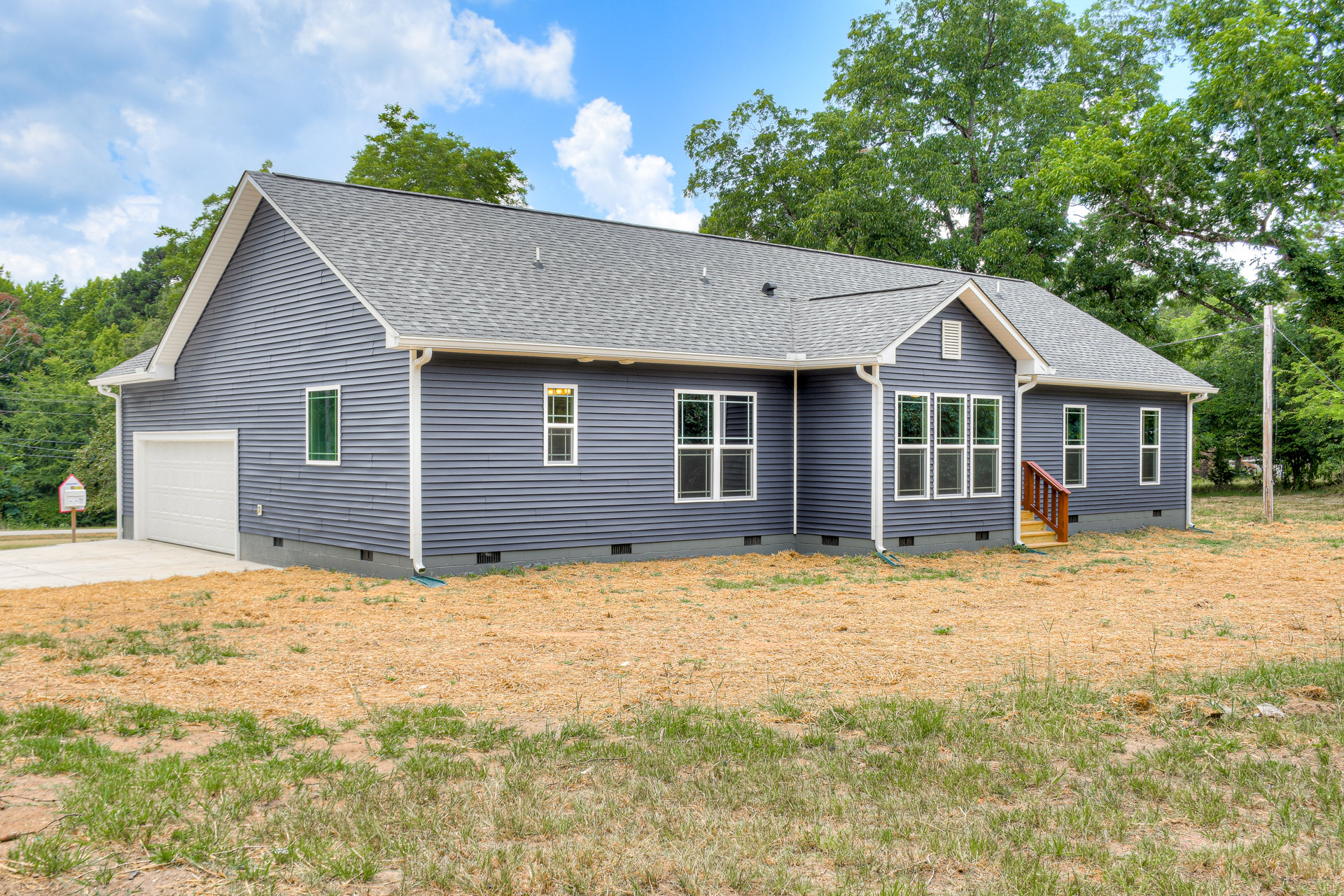 Two-story house with attached garage, white-framed windows, wooden siding, grass lawn, wood chip landscaping, and mature trees under a partly cloudy sky
