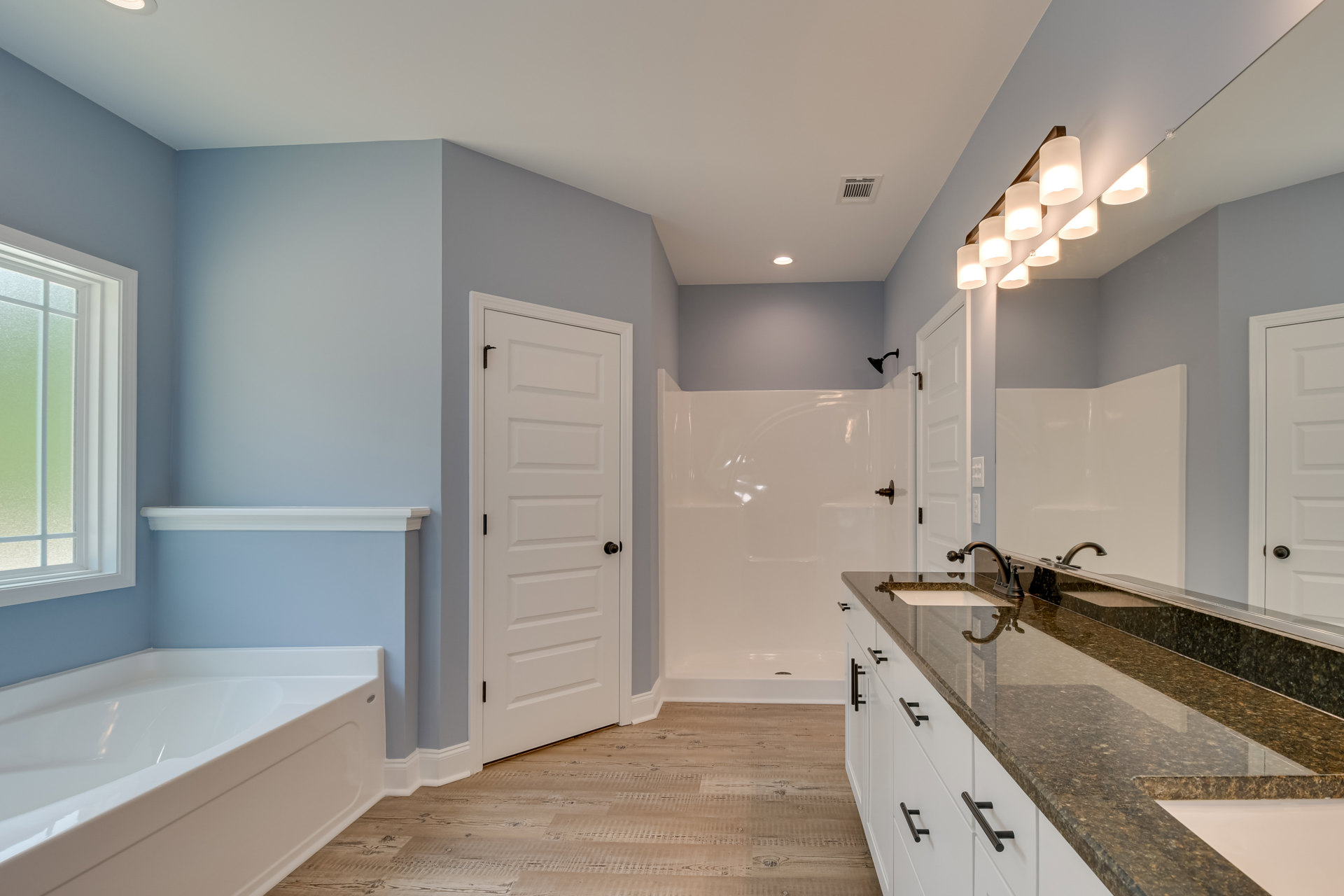 Bathroom featuring a marble countertop with white cabinets and black handles, freestanding white bathtub, glass shower enclosure, and large window providing natural light.