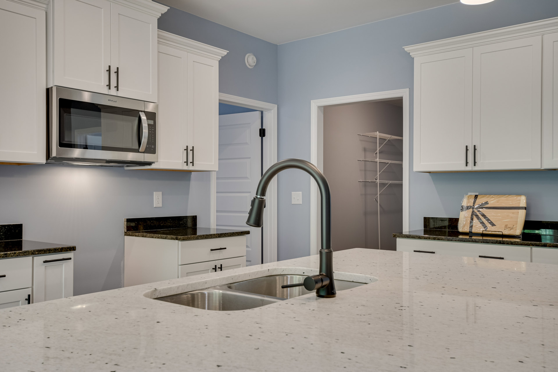 Modern kitchen with white cabinetry, black faucet and sink, open microwave above countertop, wooden board wrapped in plastic, and drawers beneath counter