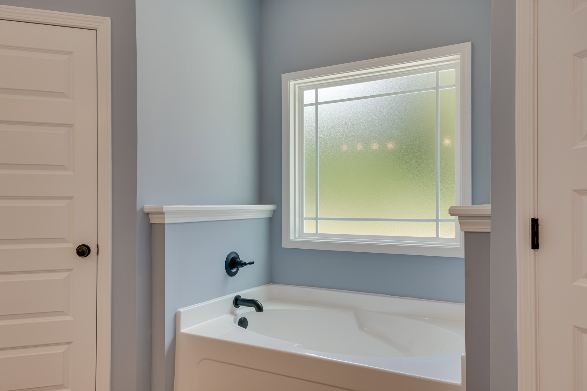 White freestanding bathtub with matte black faucet, frosted glass window, white door with silver handle, black wall switch, light gray walls