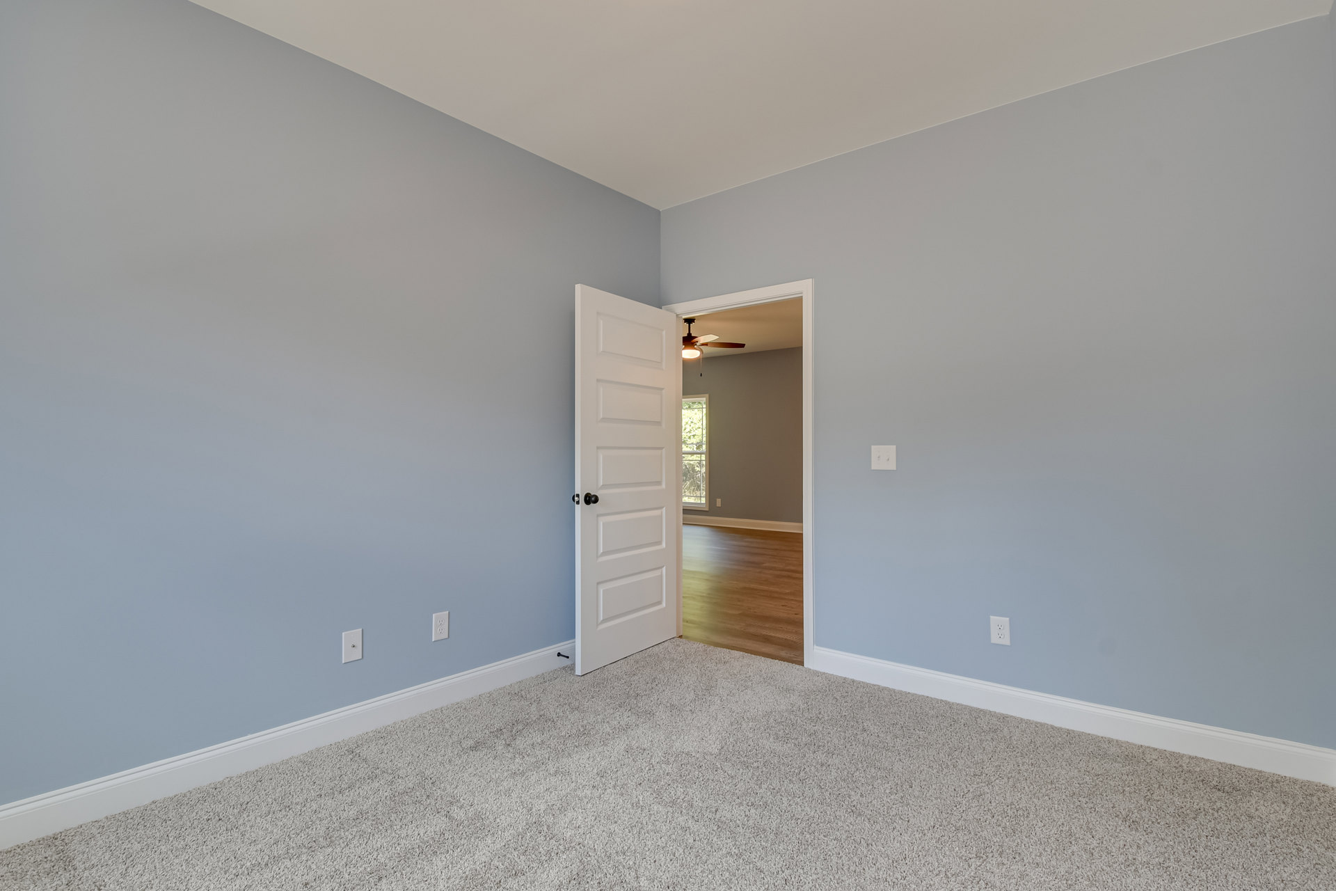 White paneled door with black knob open to a room featuring wood flooring, white area rug, plaster walls, ceiling fan with light, and visible ceiling corner.