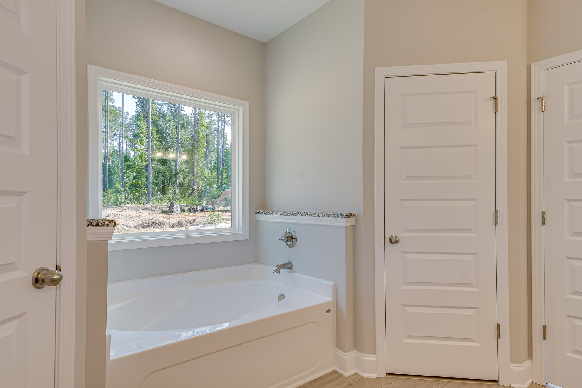 Freestanding white bathtub with chrome faucet beside window overlooking trees, white paneled door with silver knob and visible hinge, light-colored walls