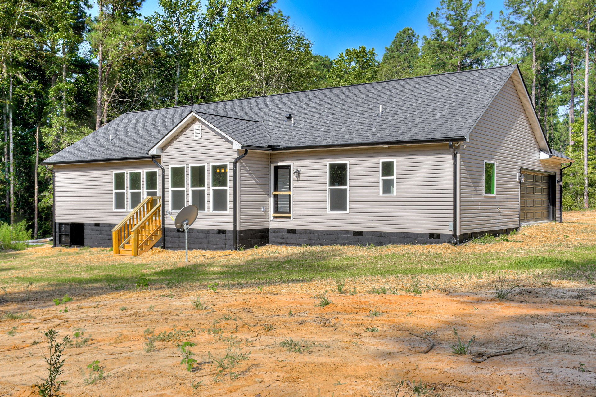 Two-story house with grey roof, white-framed windows, and wooden staircase leading to entrance; satellite dish mounted on pole beside exterior, surrounded by grass and trees.