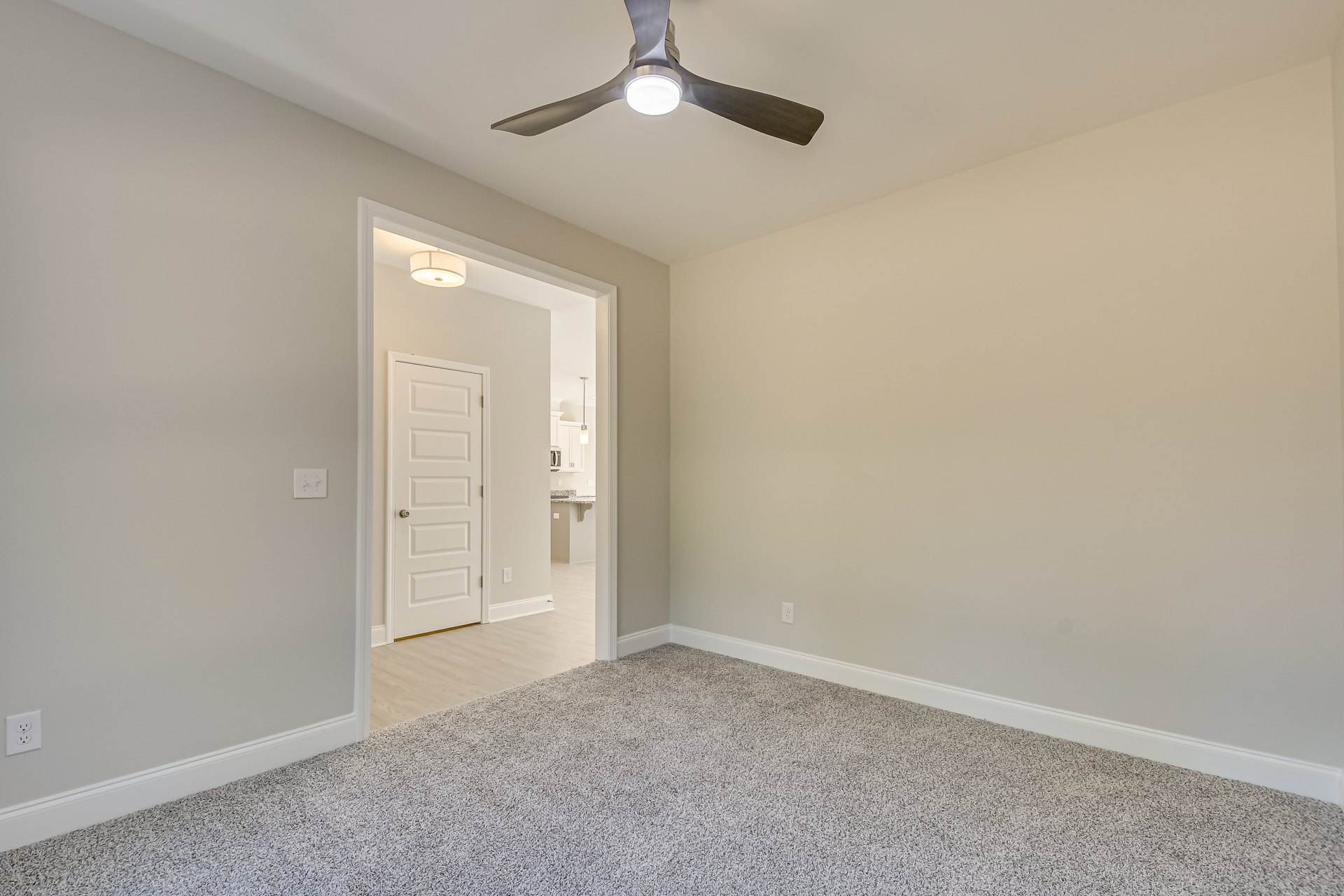 Carpeted room with white walls, ceiling fan with light fixture, white door with silver knob, wall clock above baseboard.