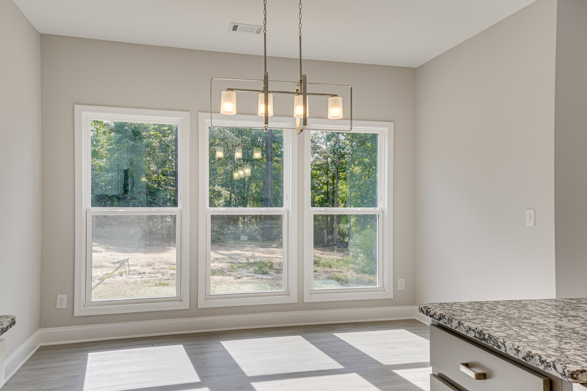 Chandelier hanging from white ceiling in bright room with large windows, white floors, and tree views outside; countertop with metal handle visible in foreground