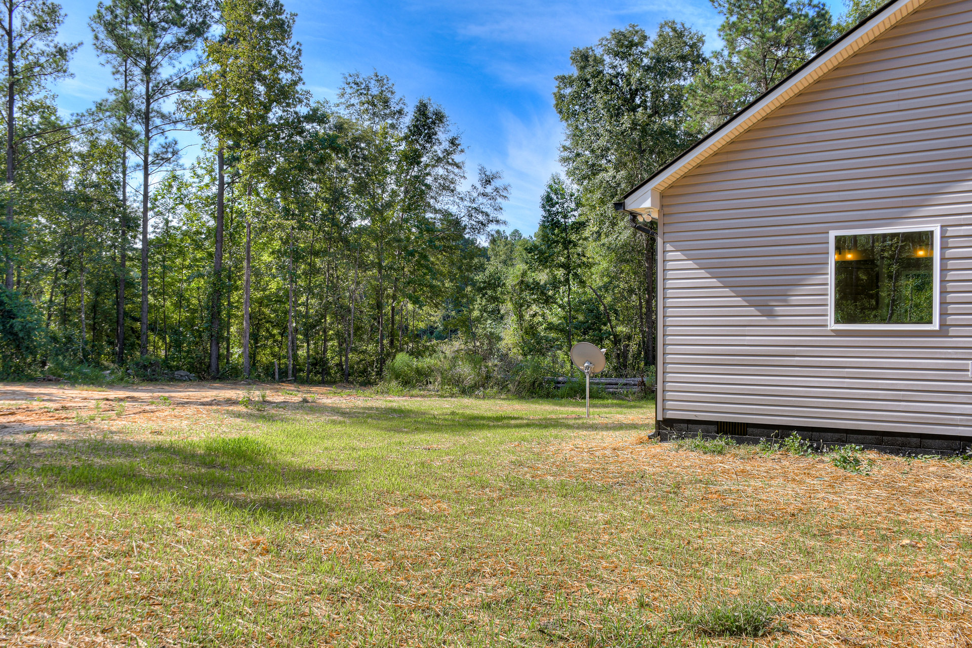 Two-story house with white siding, large windows, basketball hoop on concrete pad in grassy backyard, mature trees, satellite dish on metal pole near wooded area, partly cloudy sky