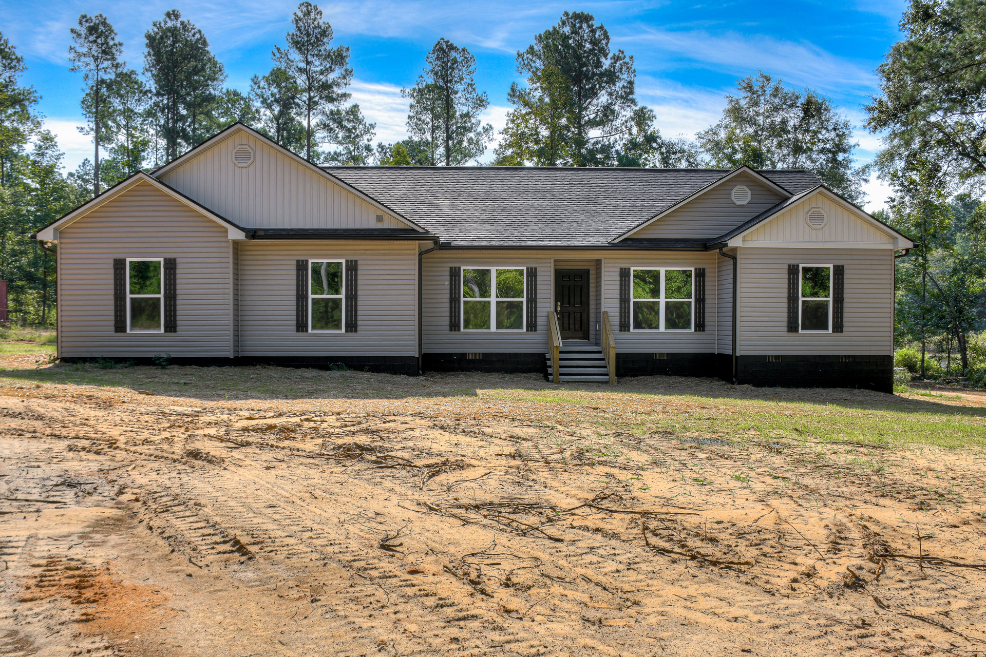 Modern cottage-style home with white-framed windows, black doors, and wooden exterior, set on a dirt lot with mature trees and cloudy sky in the background