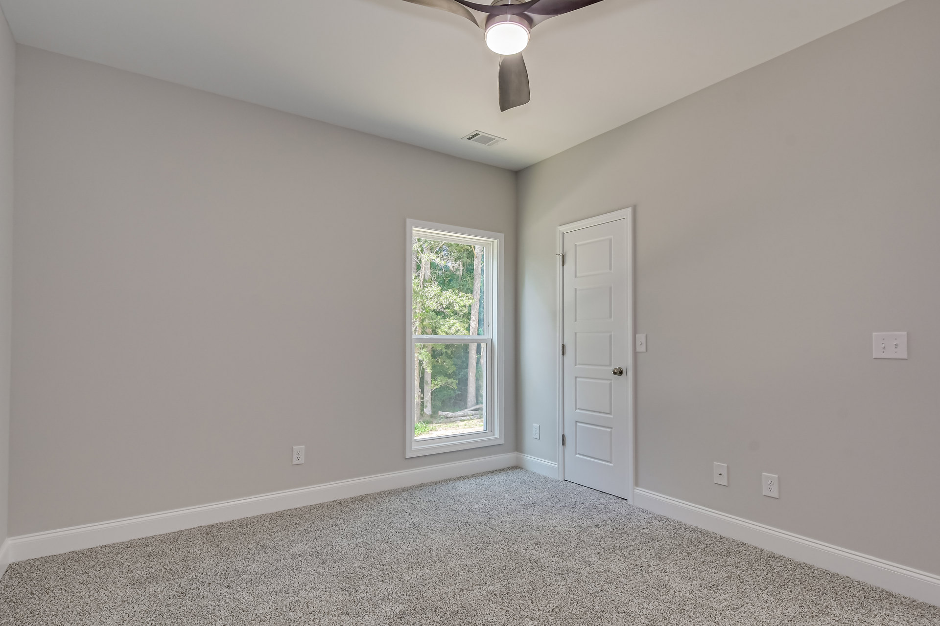 Carpeted bedroom with white trim, ceiling fan with light, large window showing trees outside, white door with silver knob