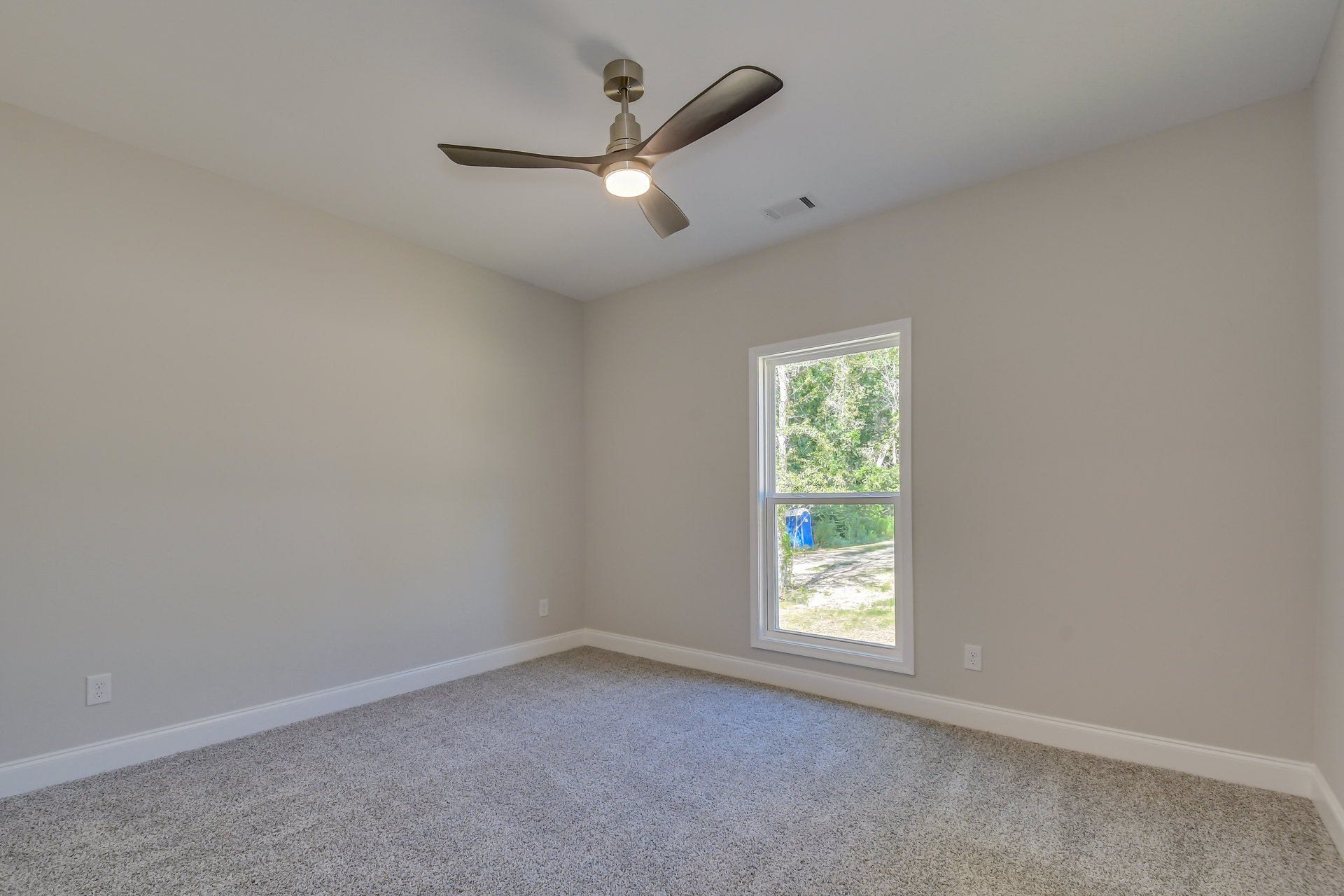 Ceiling fan with integrated light fixture mounted on white plaster ceiling, carpeted floor, window with blue container and trees visible outside, simple wall molding.