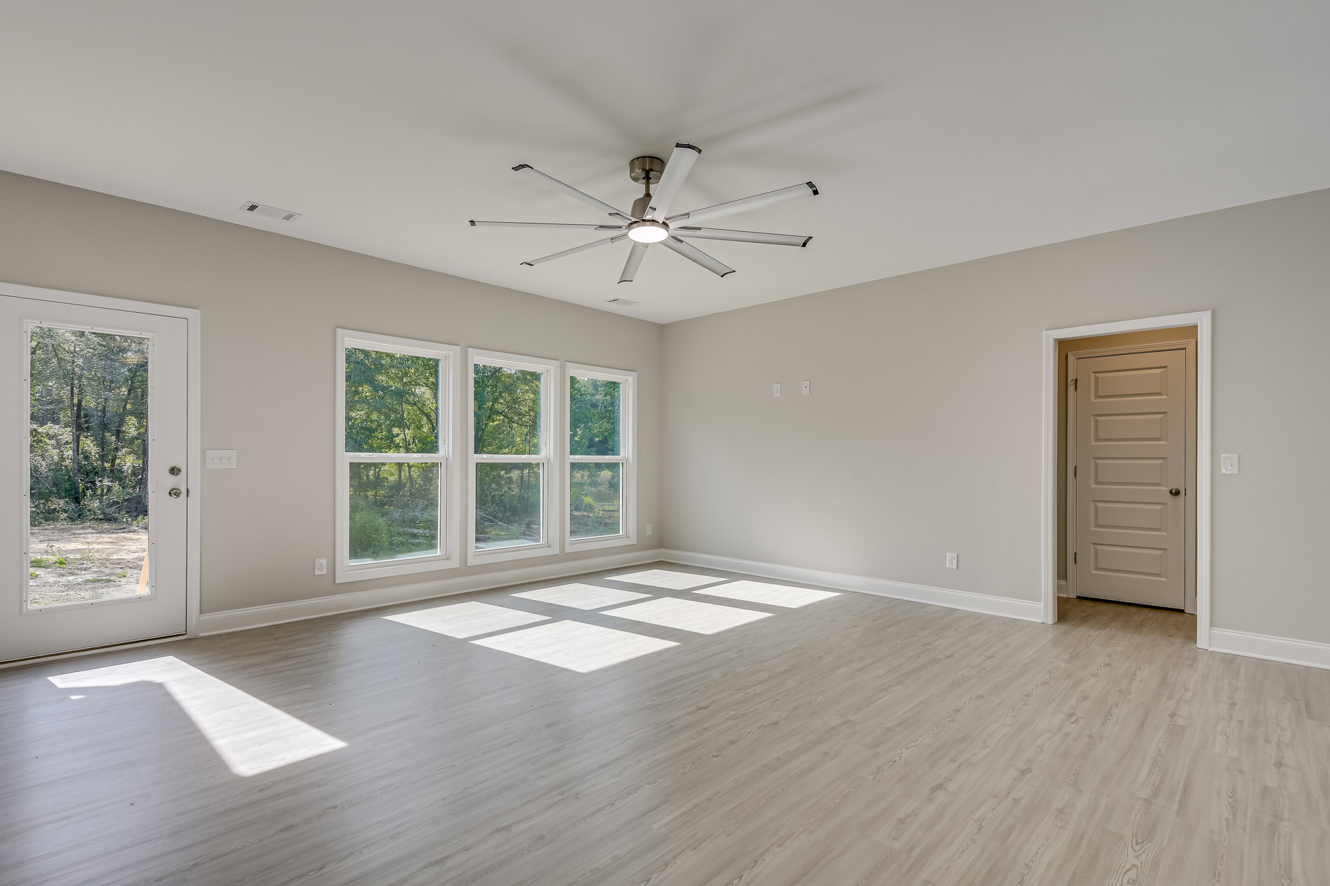 Ceiling fan with light fixture, white door with silver knob, wood flooring, large windows showing trees outside, neutral plaster walls