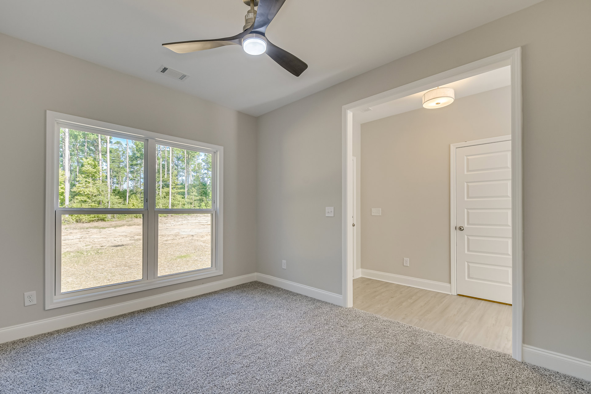 Carpeted room with white walls, ceiling fan with light fixture, single window showing trees outside, white door with silver knob