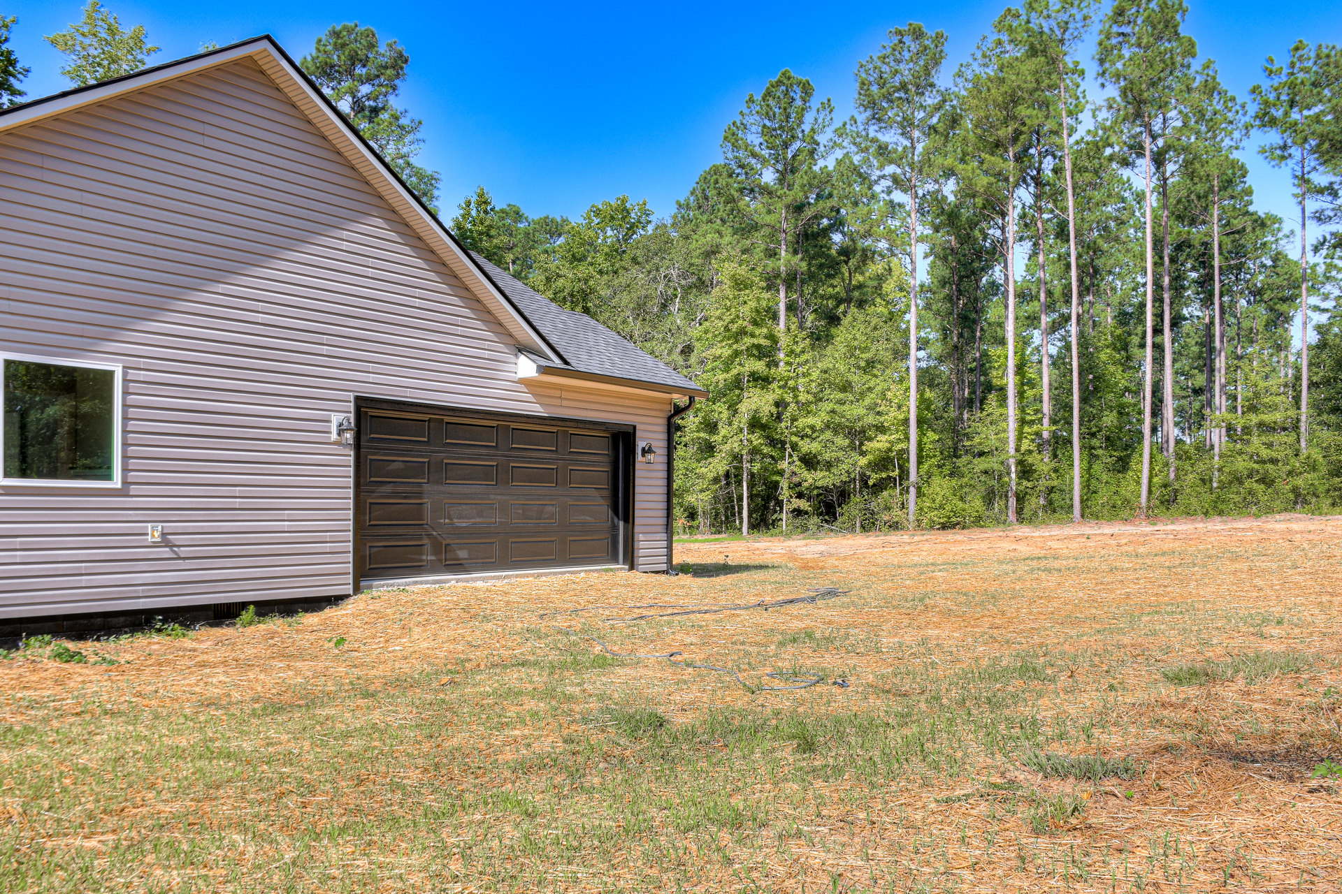 Gray garage door with upper windows, white-framed windows on beige siding, manicured grass lawn, tall trees behind house