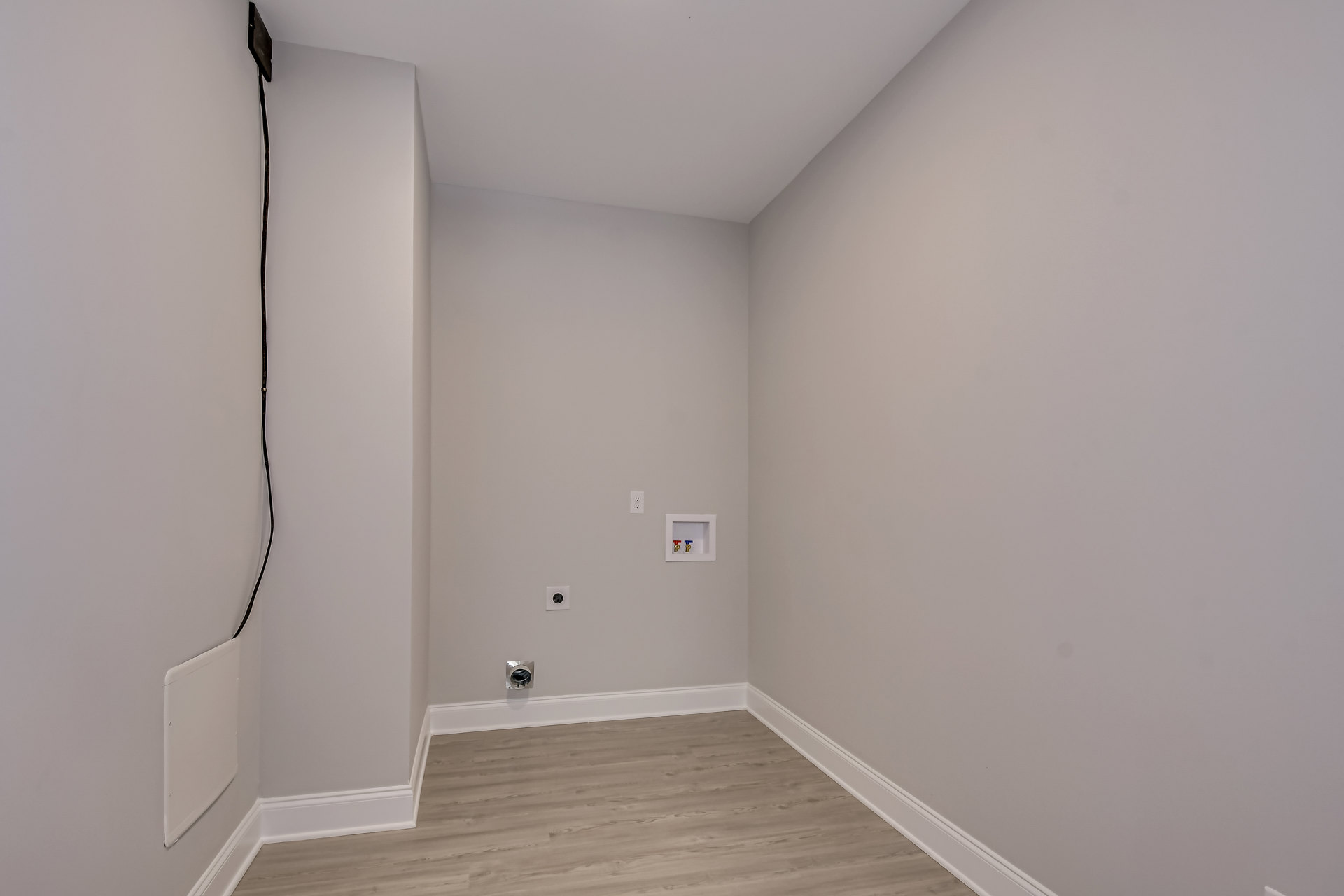 Wood floor with white baseboard trim, white plaster walls, ceiling-mounted light fixture, and a white square panel with red and blue valves.