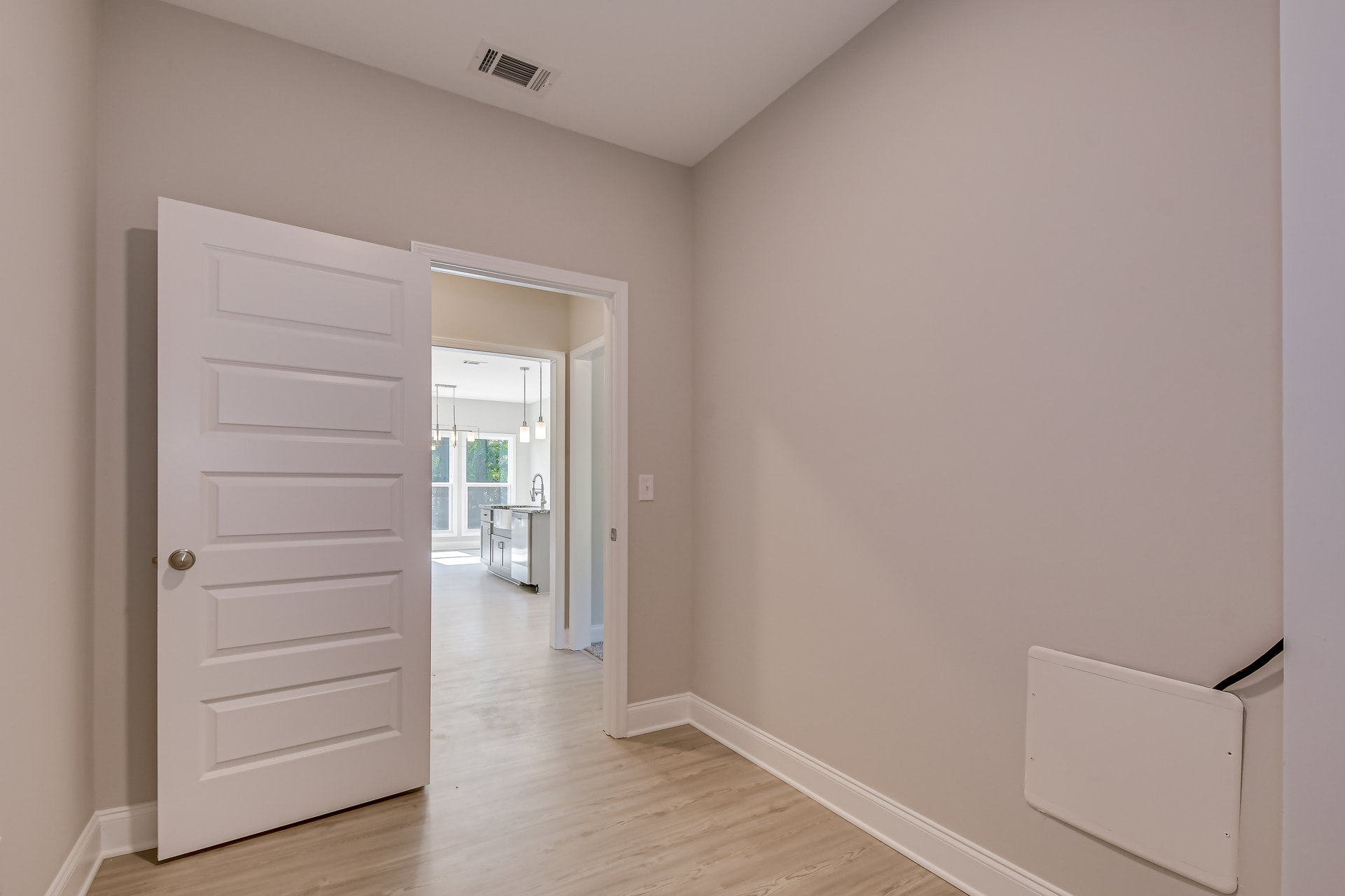 Open white door with silver handle leading to room with wood flooring, white walls, ceiling vent, and visible kitchen sink