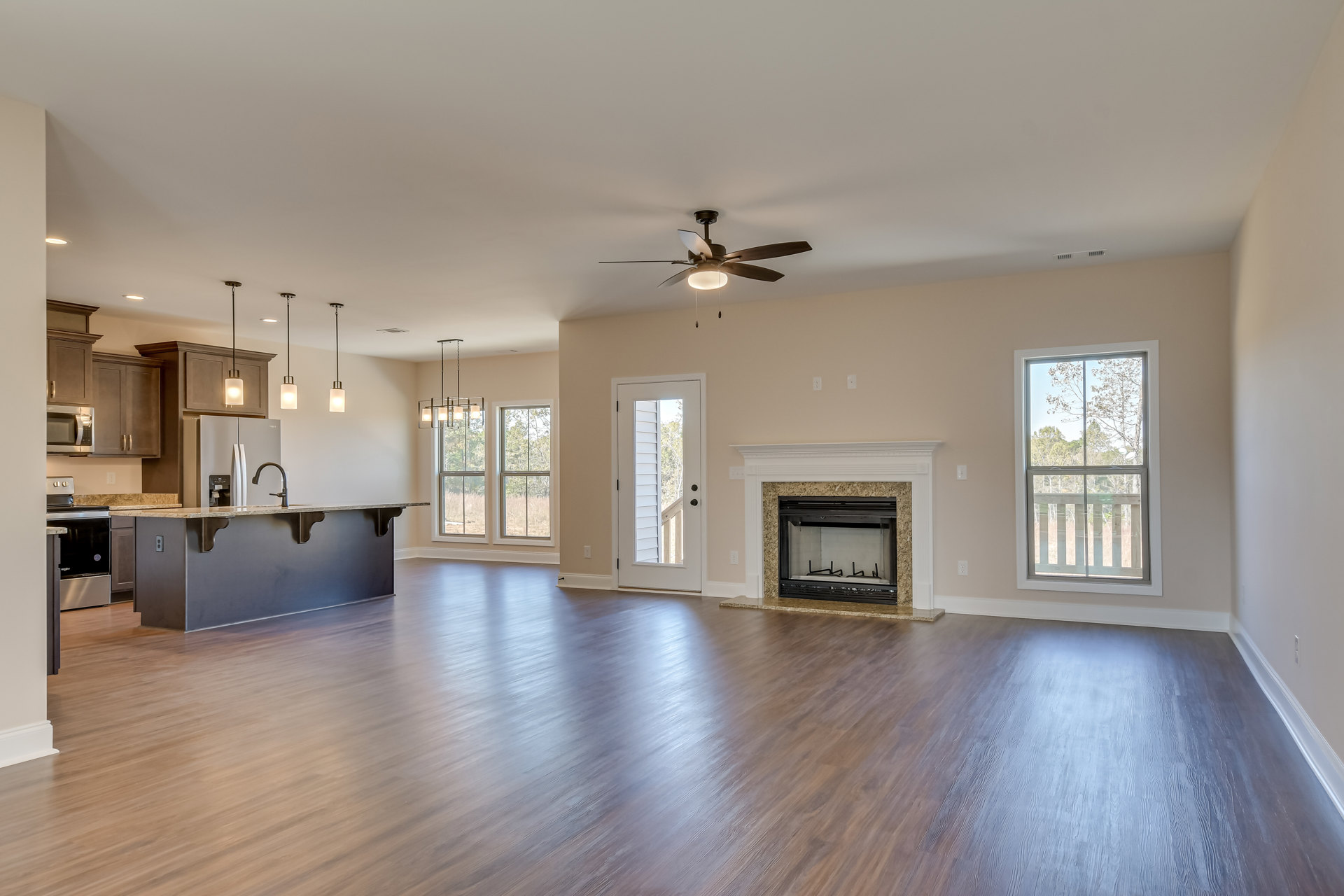 Spacious living room featuring hardwood floors, central glass-door fireplace, open kitchen area, ceiling fan with light, large windows and door offering views of trees outside