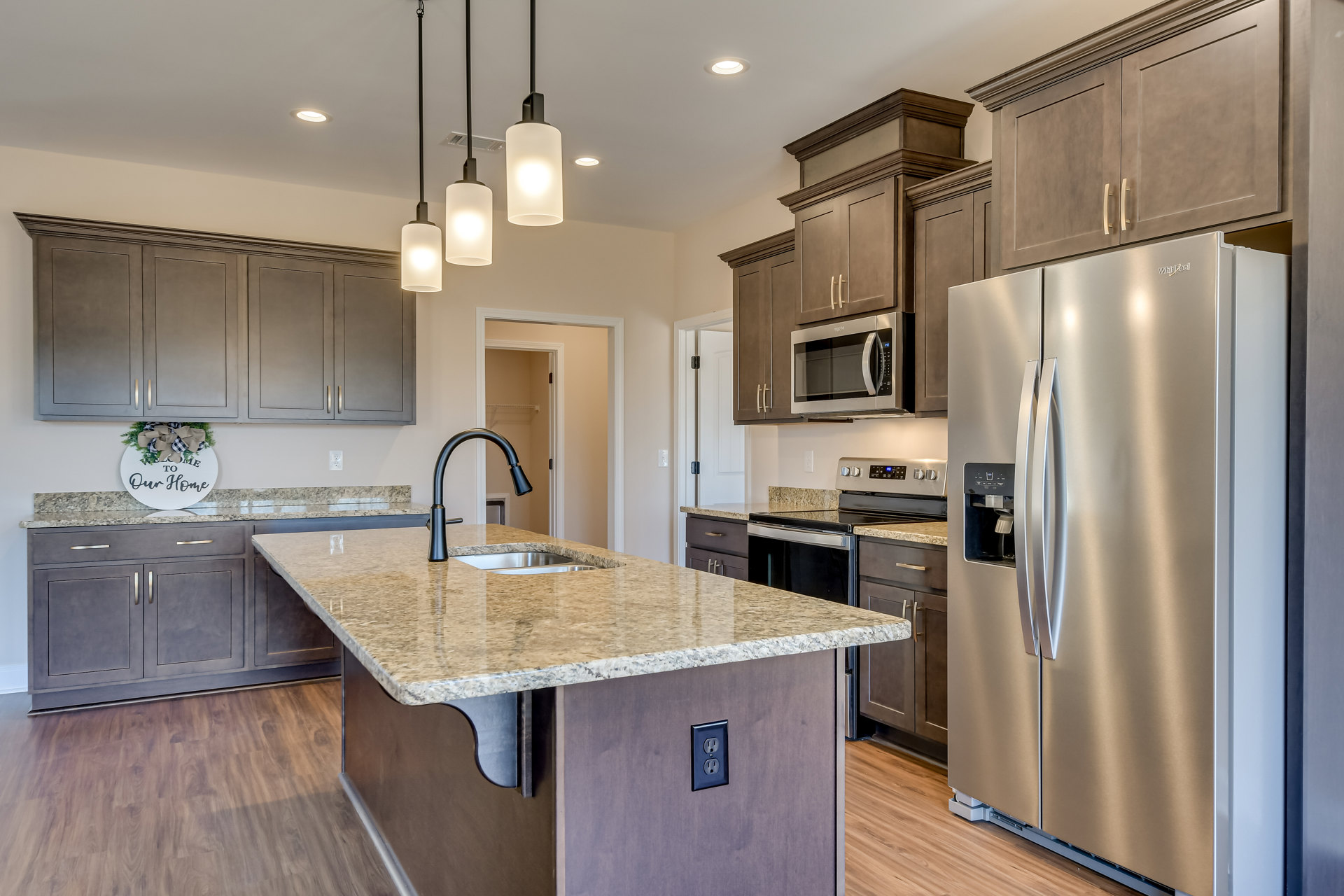 Granite kitchen island with built-in sink, stainless steel refrigerator, microwave, and cabinetry; wall outlet and decorative sign with bow visible