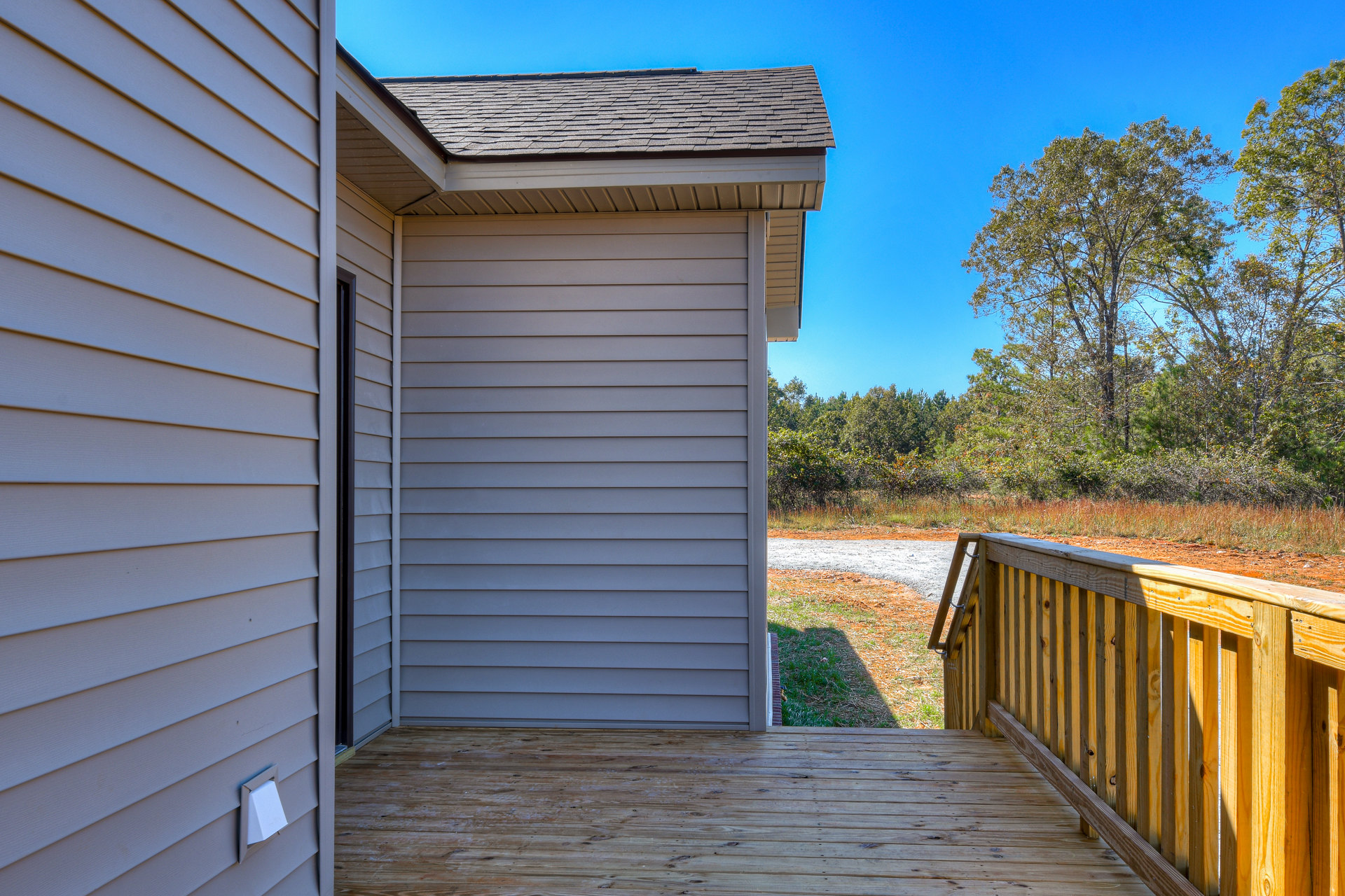 Wooden deck with horizontal railing, white electrical box mounted on siding, grassy yard, glass door, and backdrop of mature trees under blue sky.