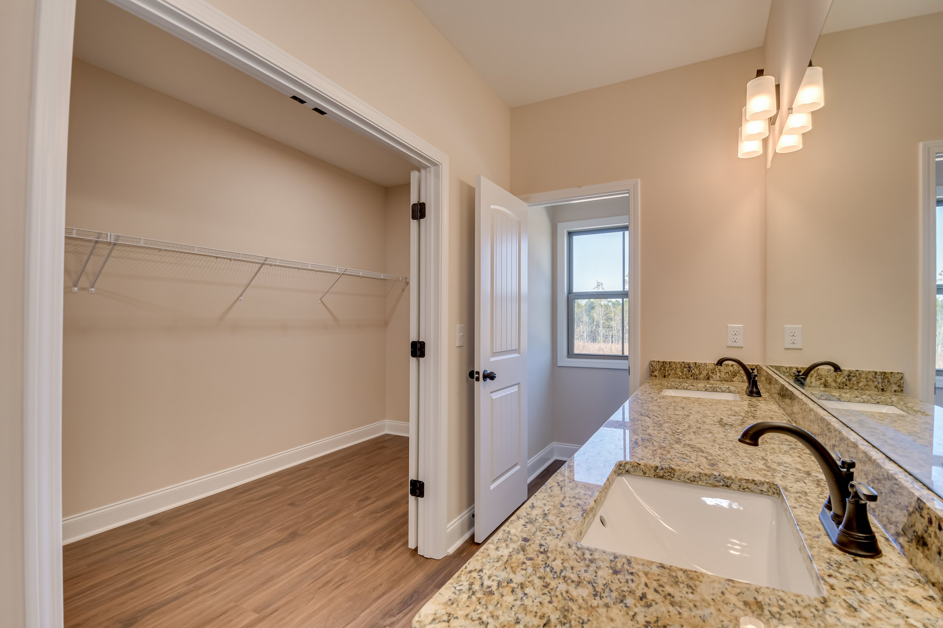 Bathroom featuring marble countertops, wood flooring, under-mount sink with chrome faucet, wall-mounted lamps above mirror, window overlooking trees, and adjacent closet with