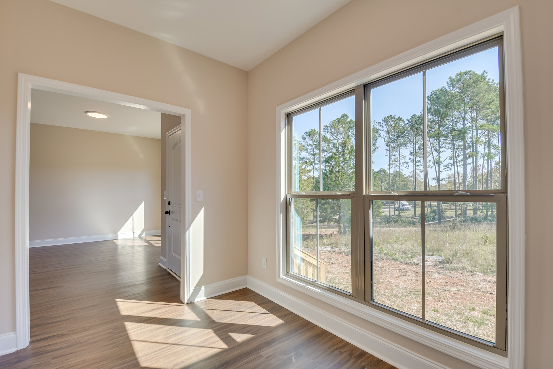 Spacious room featuring wide plank wood flooring, large picture window with views of trees and open field, white walls, and adjacent door
