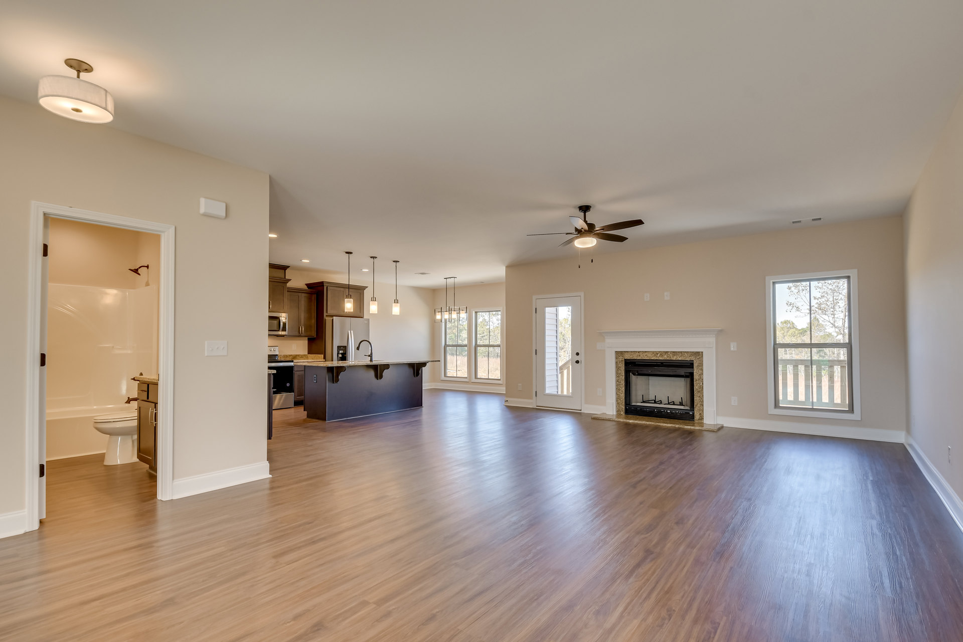 Spacious open floor plan featuring a black fireplace, hardwood flooring, white doors, and a kitchen area; large windows reveal tree views outside.