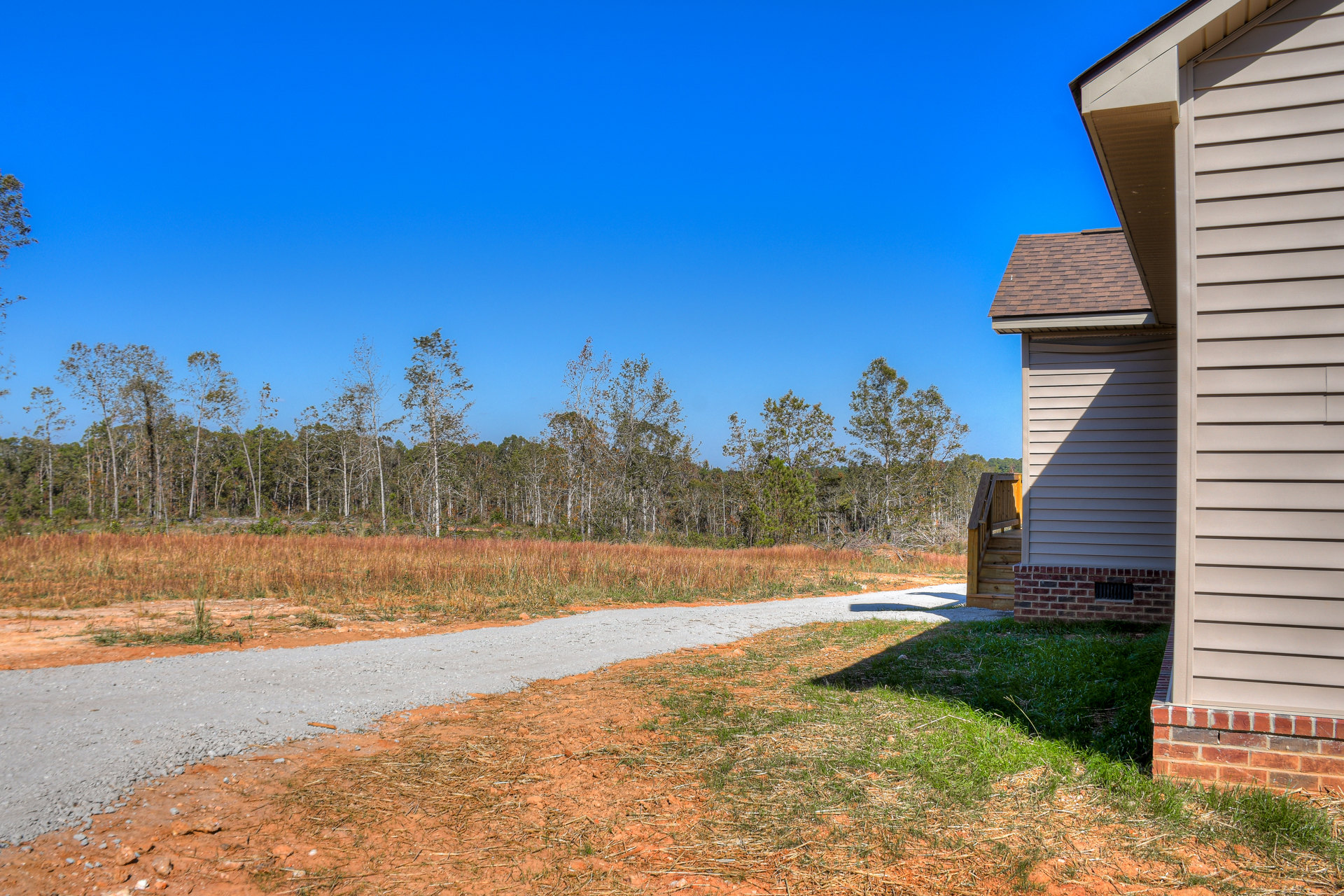 Two-story home with light siding, gabled roof, and front path bordered by grass, mature trees in the background, blue sky overhead
