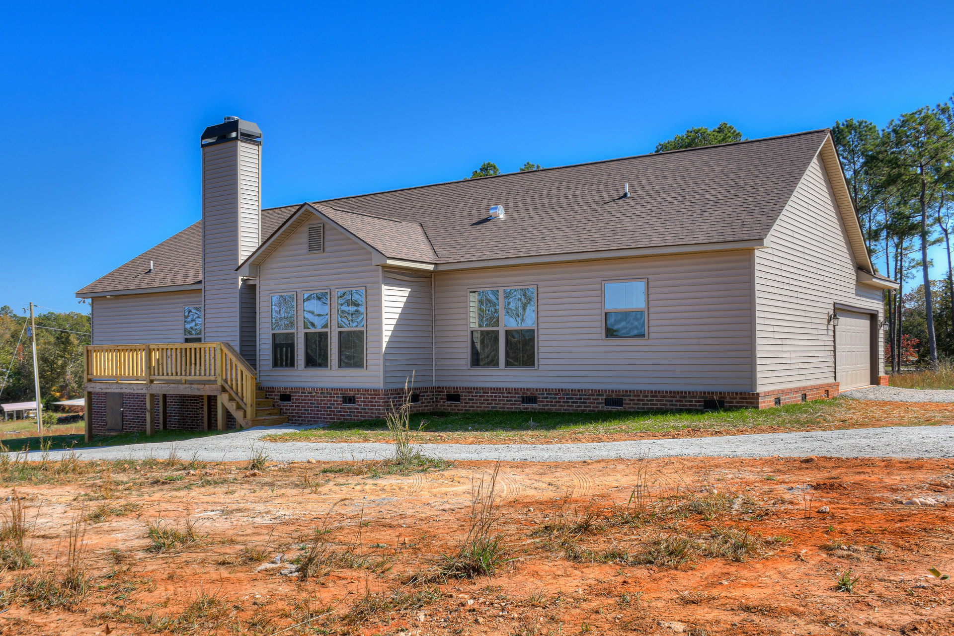 Wood deck attached to a house with brick wall, large windows reflecting nearby trees, pitched roof, dirt road bordered by grass, surrounded by outdoor plants and trees