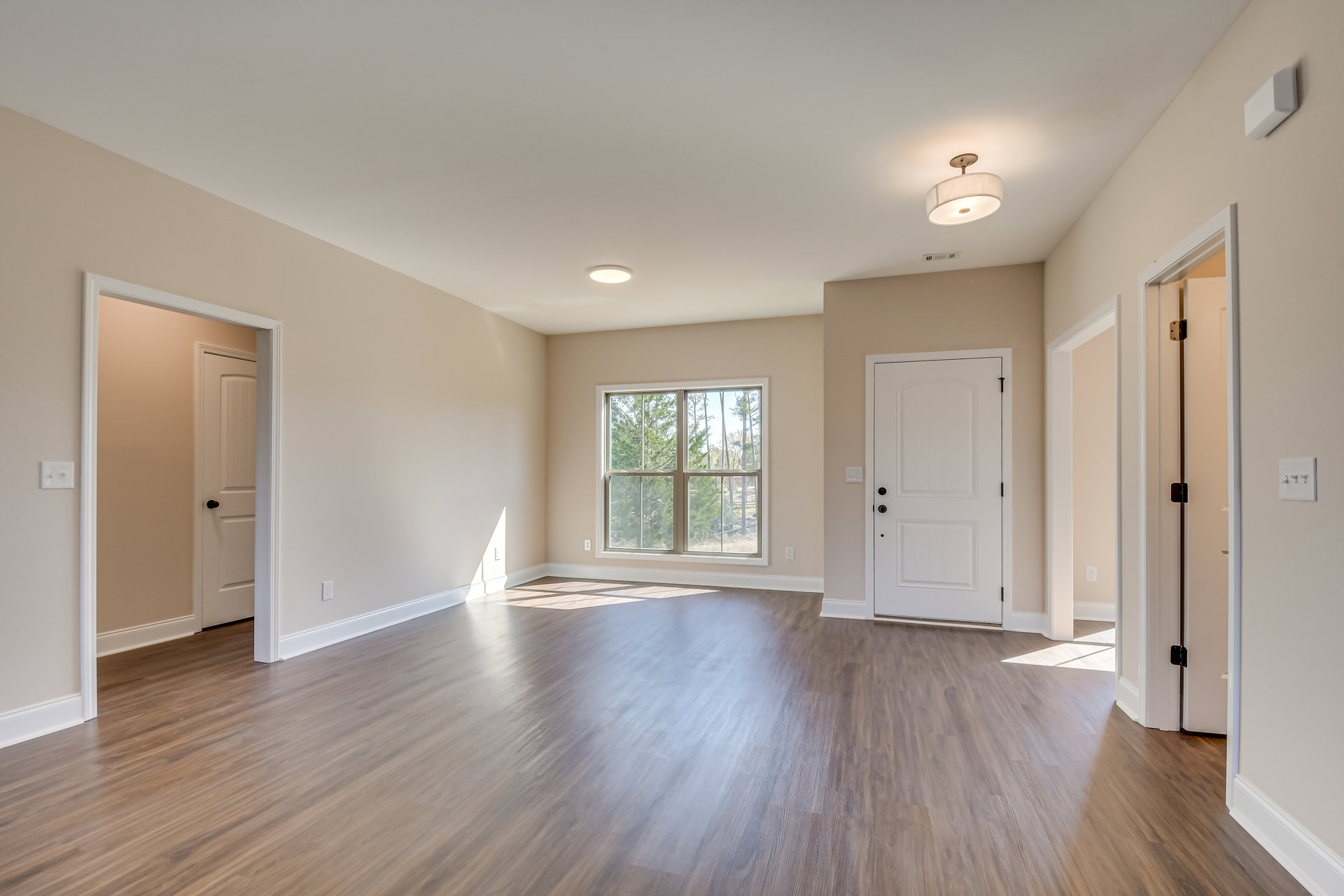 Wood flooring in a bright room with a large window overlooking trees, white door with black knobs, and ceiling light fixture with white shade