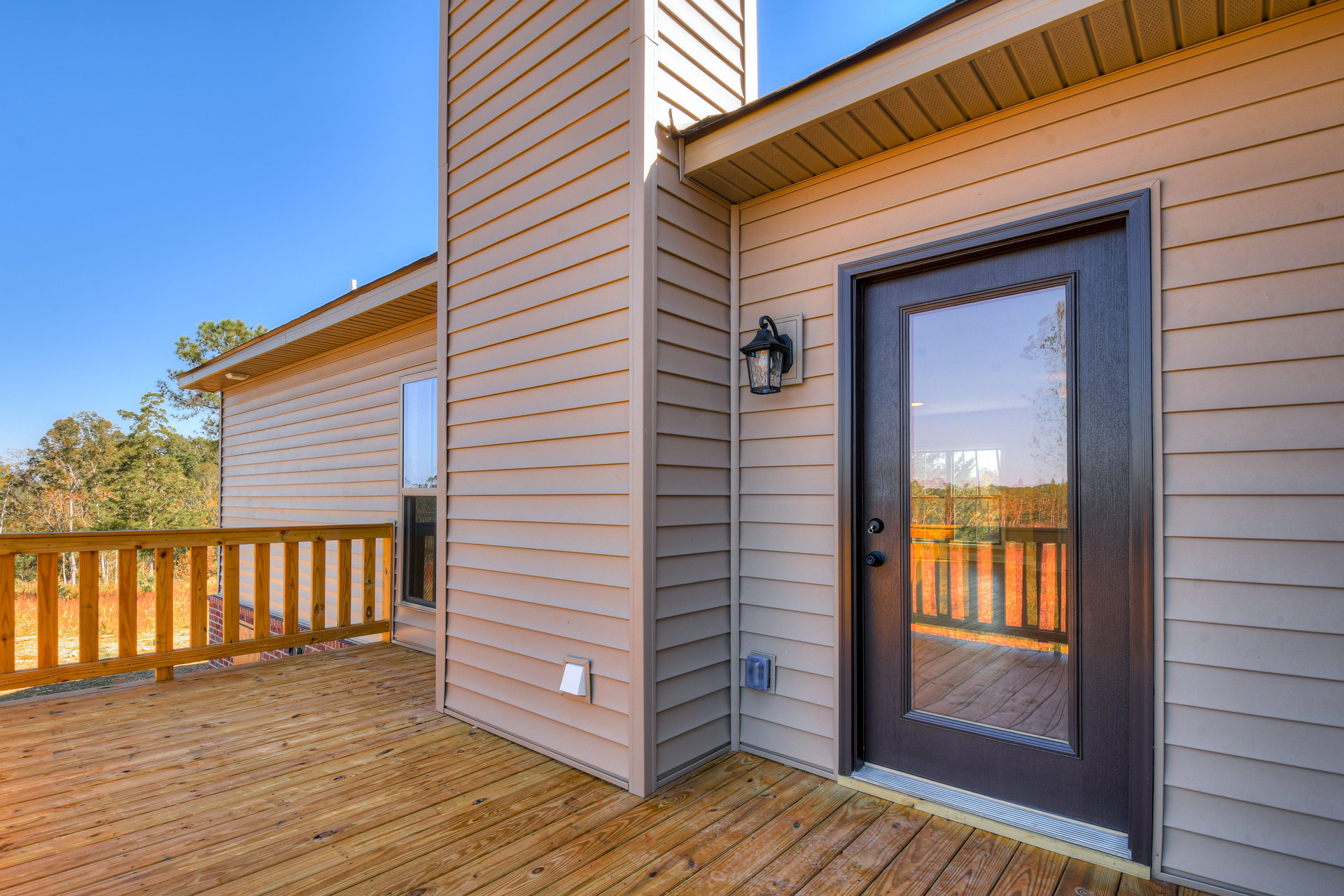 Wooden deck with horizontal railing, glass door reflecting trees, adjacent window with glass panel, exterior siding in neutral color, porch lamp mounted near entry