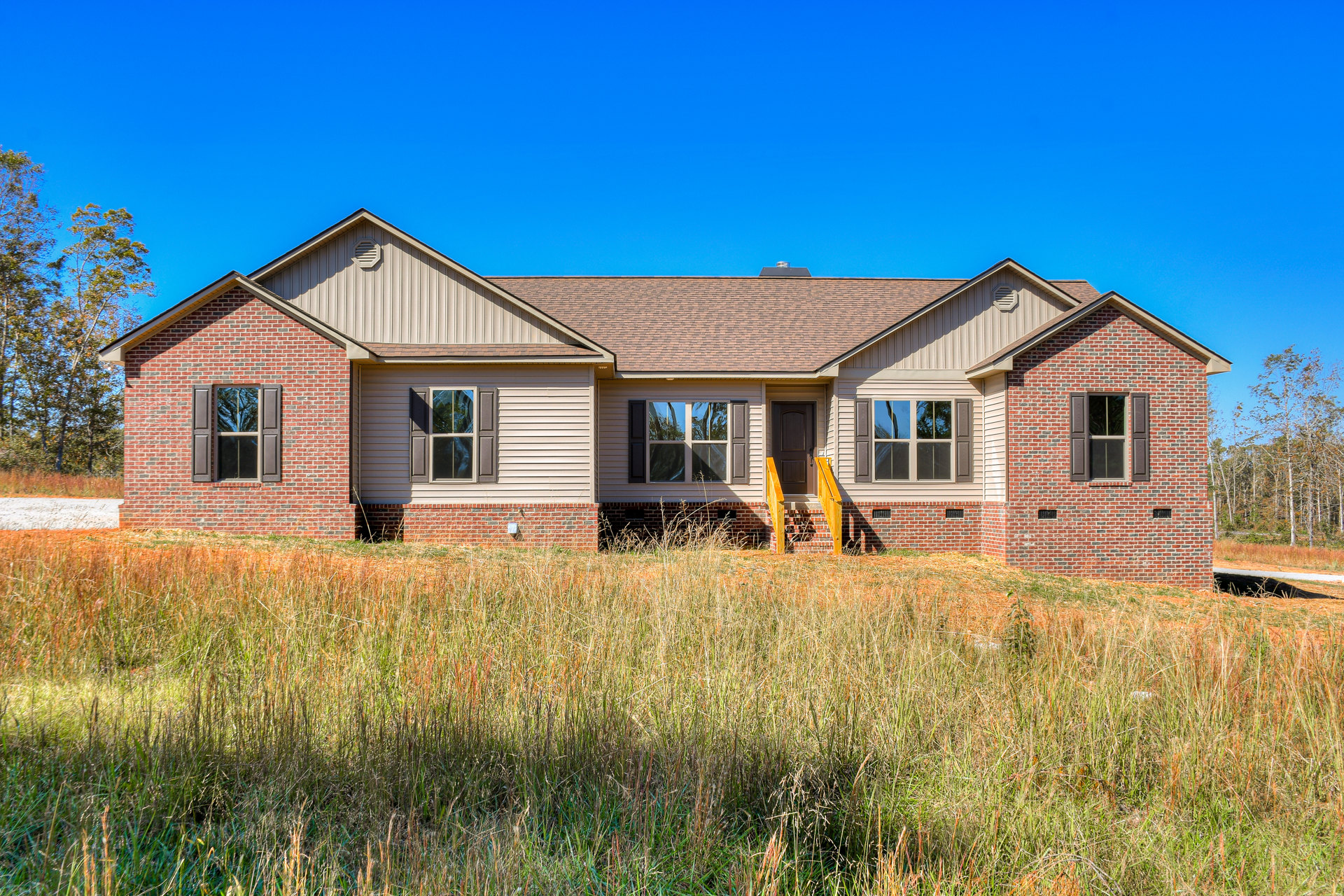 Brick house with brown roof, tall grass in foreground, multiple windows reflecting nearby trees, open grassy field surrounding exterior