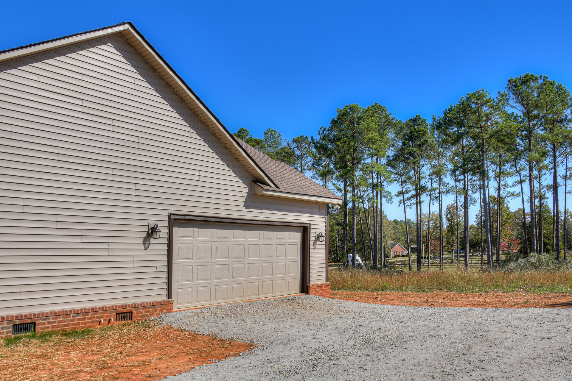Two-story house with white roof, brown-trimmed garage door, light siding, gravel driveway, and tall trees in the background under blue sky
