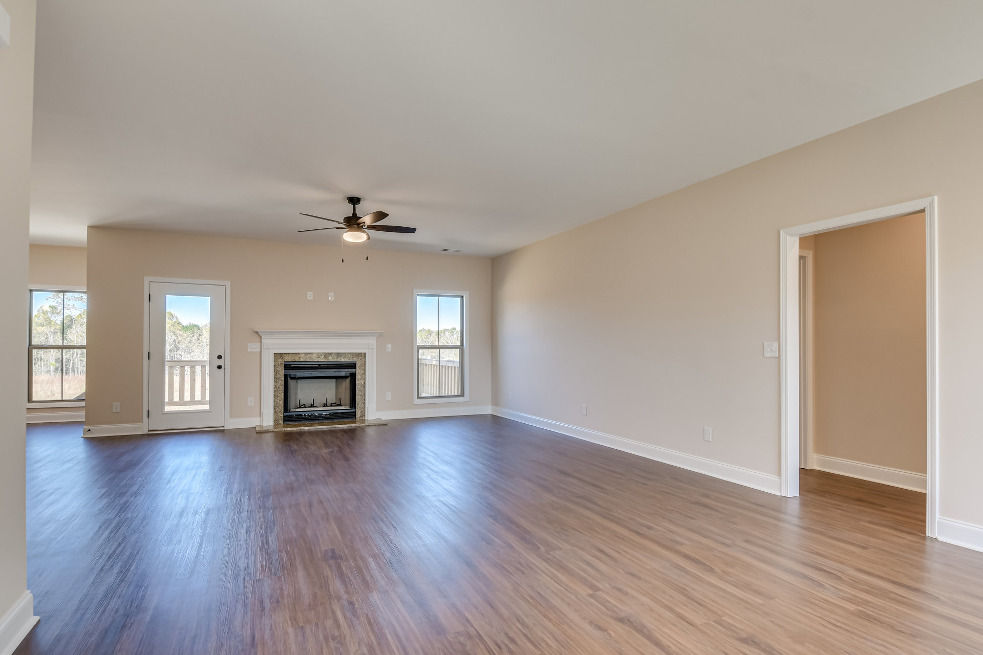 Living room with wood flooring, black-framed fireplace, ceiling fan with light, glass door with railing, large window showing trees outside