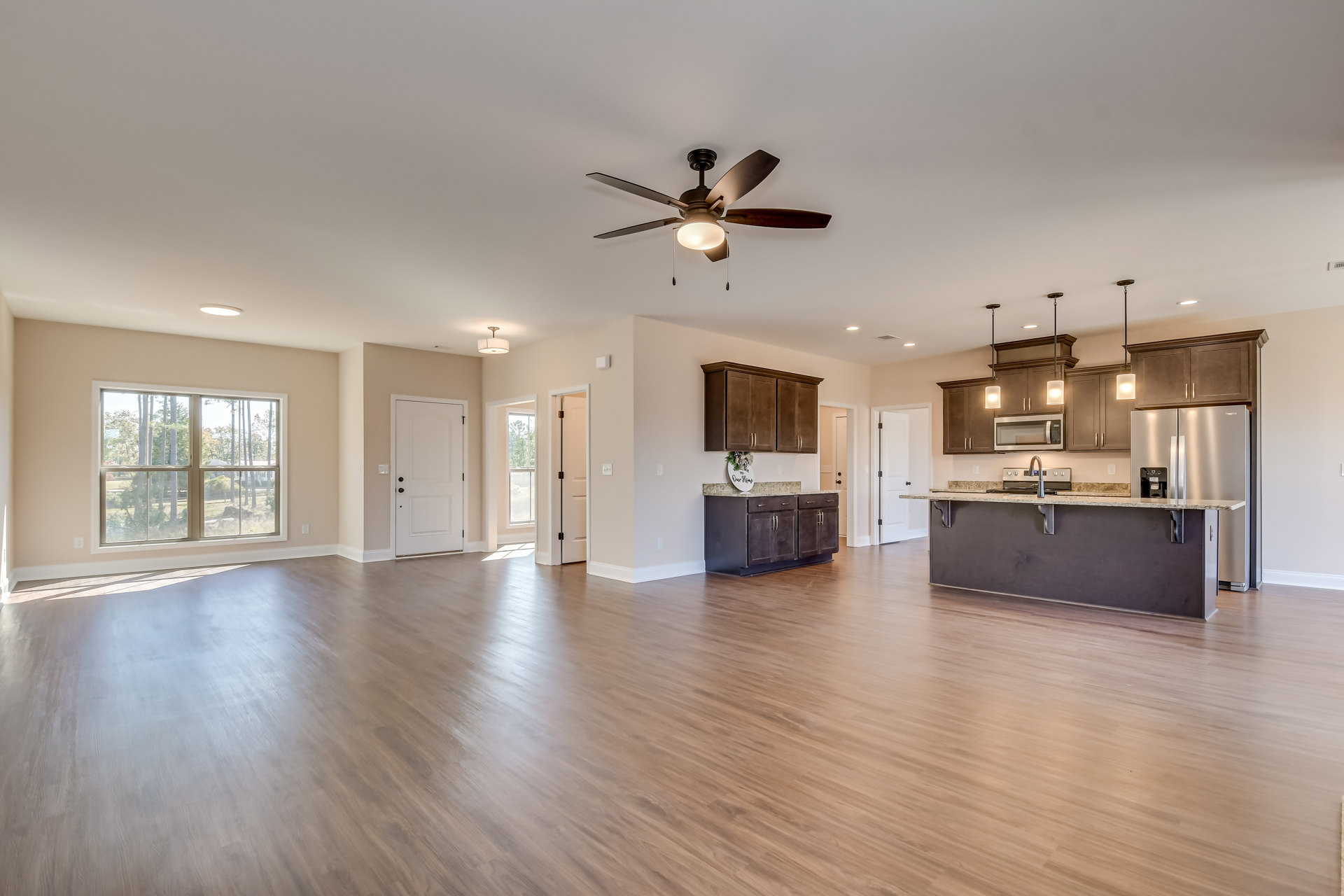 Spacious open floor plan featuring wood laminate flooring, white walls, kitchen island with white cabinetry, ceiling fan with light, dining area, large window overlooking trees