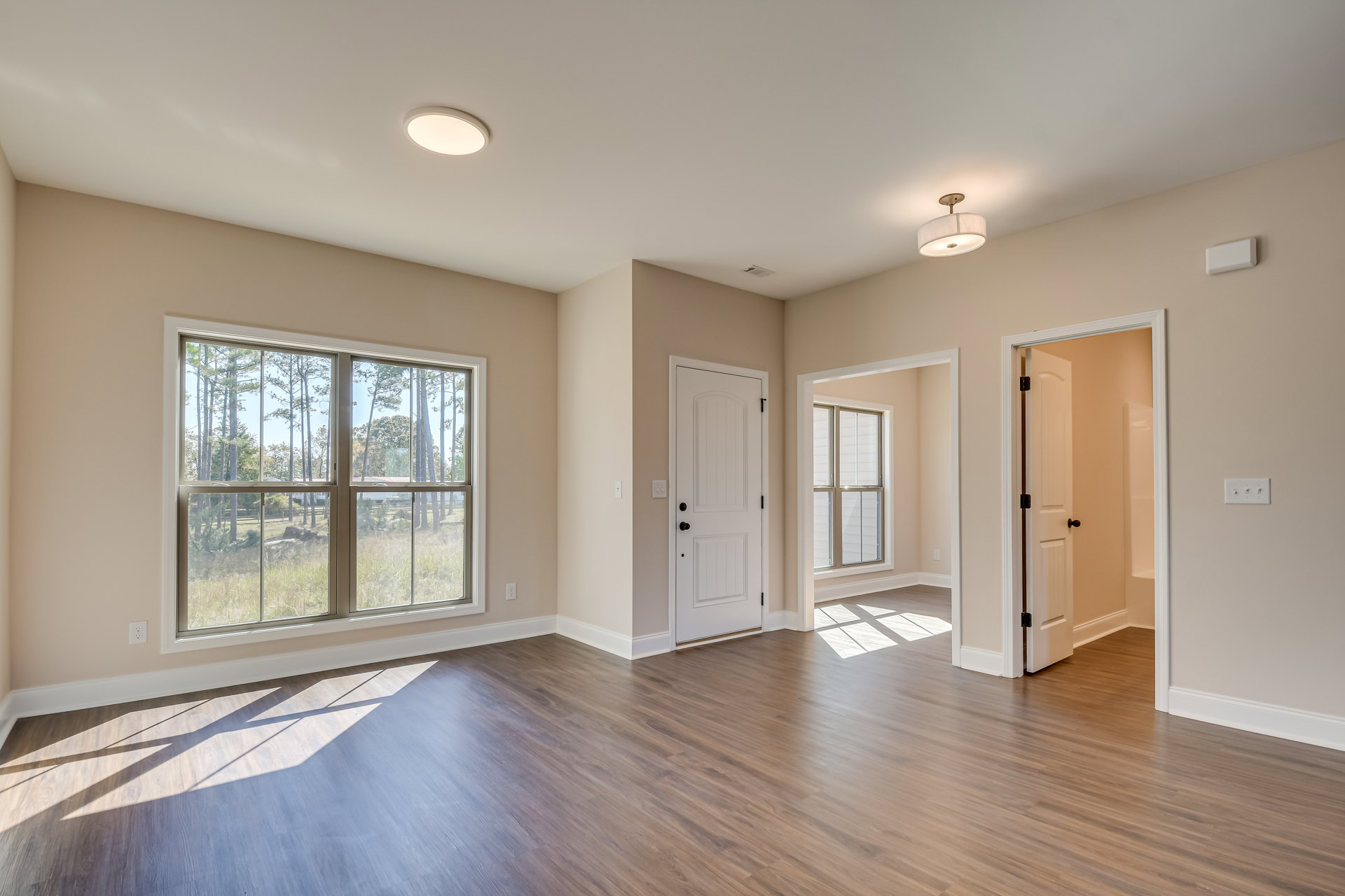 Wood flooring with white baseboard trim, large window overlooking trees, white door with black hardware, ceiling light fixture, white wall-mounted thermostat