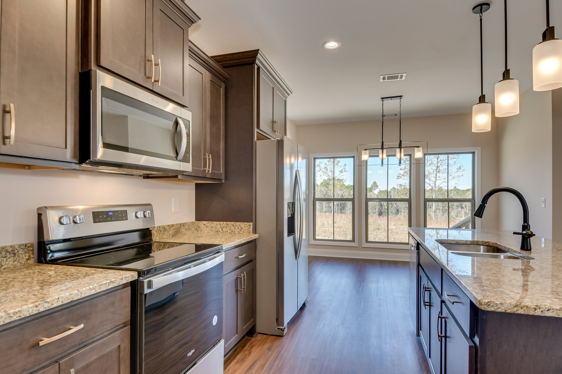Kitchen with wood flooring, white cabinetry, stainless steel microwave and stove, white countertops with black accent line, window overlooking trees, and a pendant light fixture