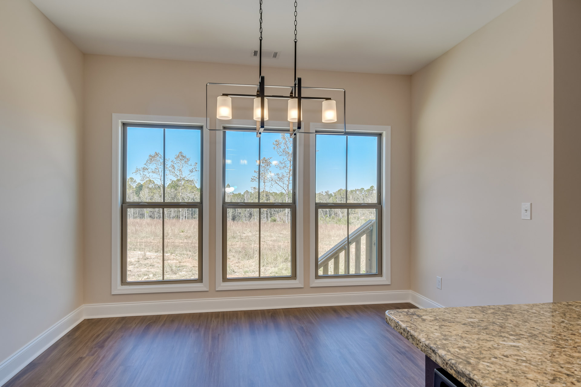 Kitchen with marble island and countertops, wood flooring, white baseboards, large window with view of trees, modern light fixture.