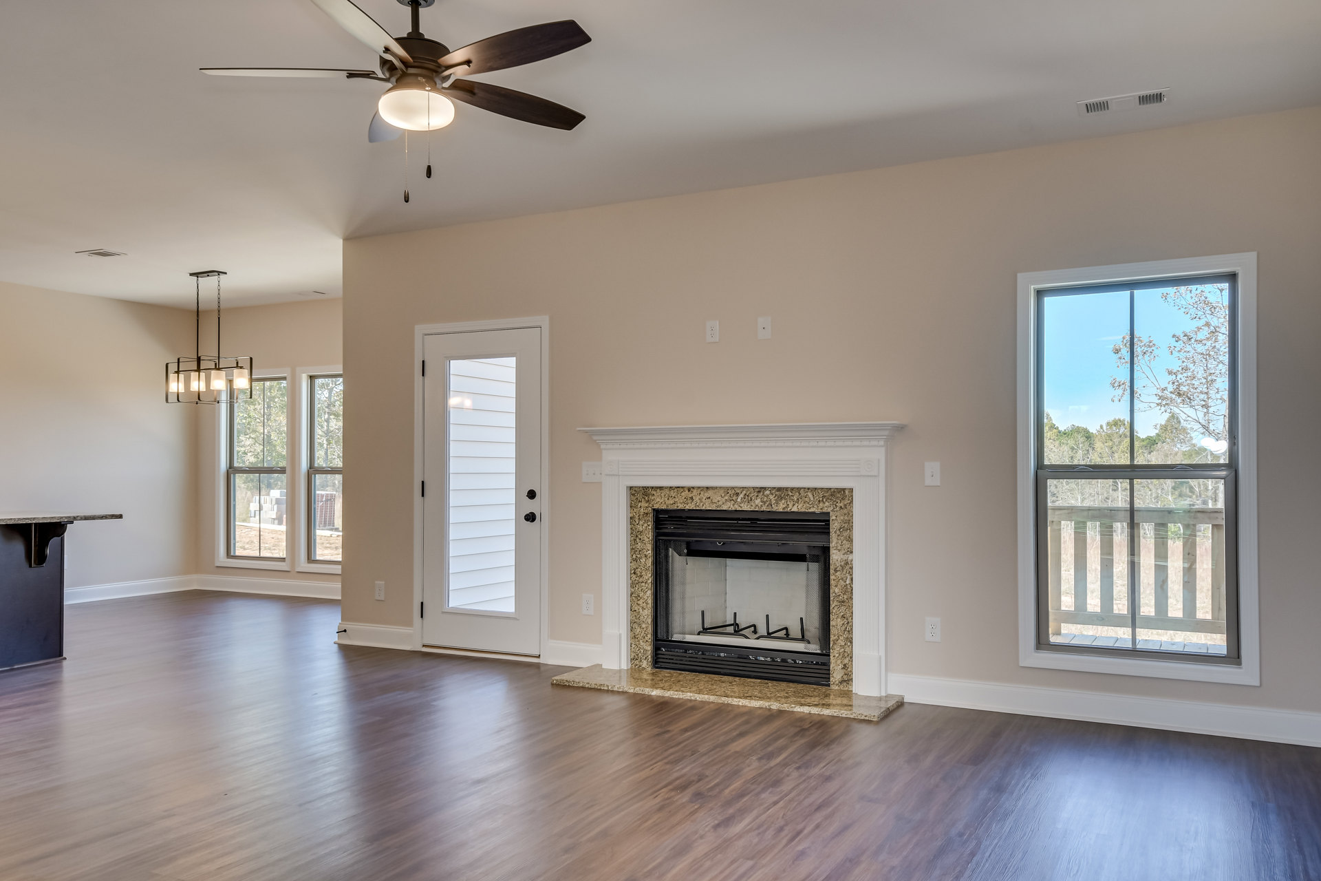 Living room with wood flooring, central fireplace featuring a mesh screen, ceiling fan with light fixture, white door with black knobs, large window overlooking trees