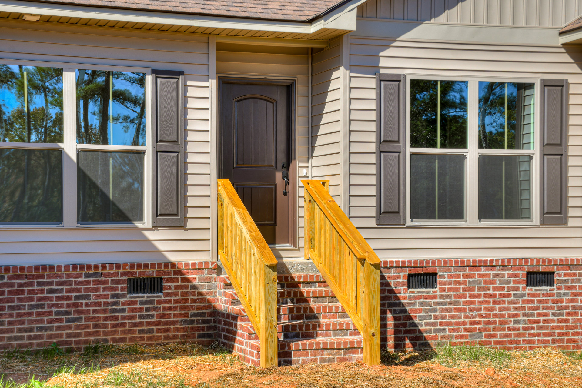 Two-story house with light siding, brick staircase leading to wooden railing, grassy lawn, large windows with screens, and wooden front door with metal handle