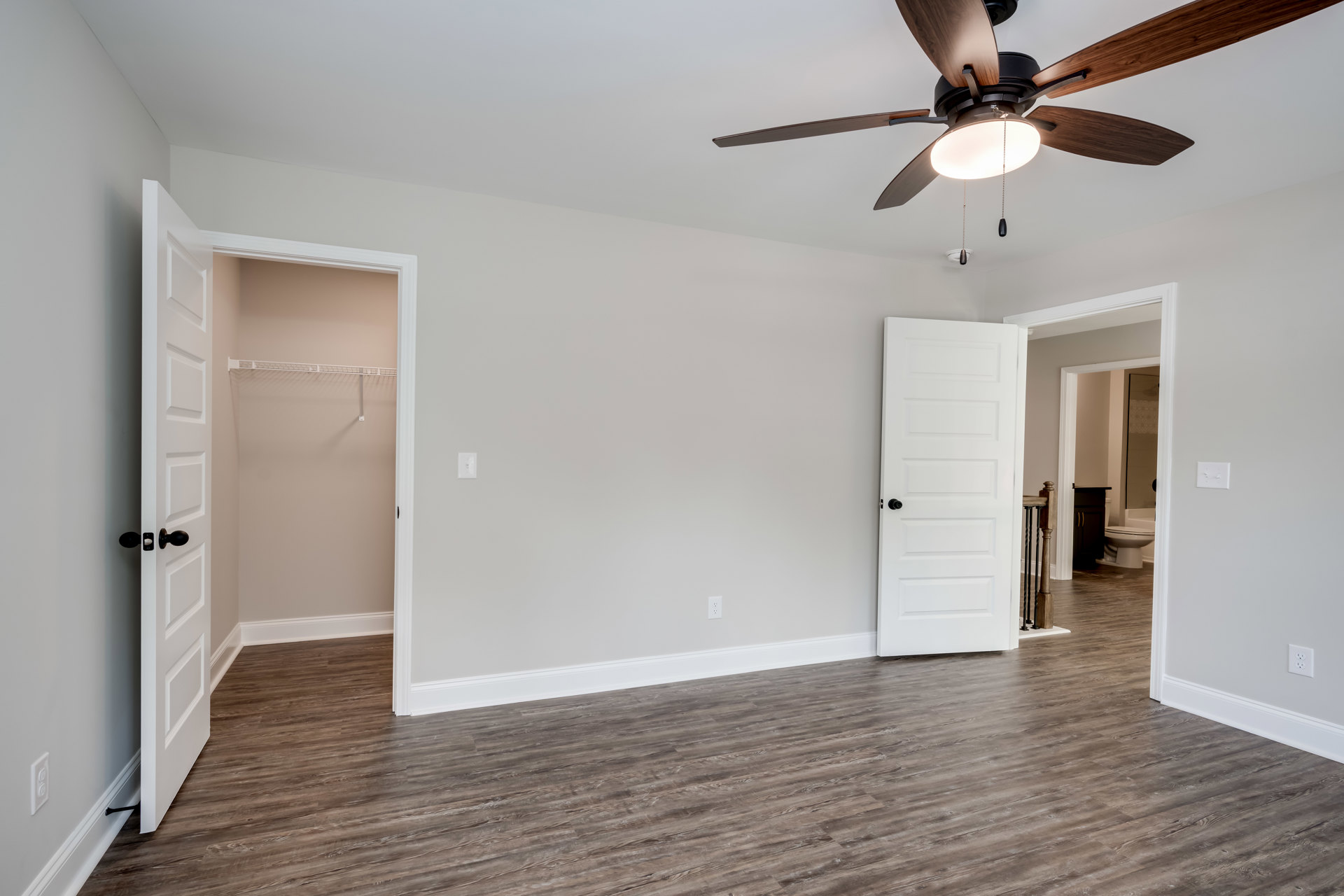 Bedroom with wood flooring, white walls, ceiling fan with light fixture, white closet door featuring black knob, electrical outlet visible near baseboard