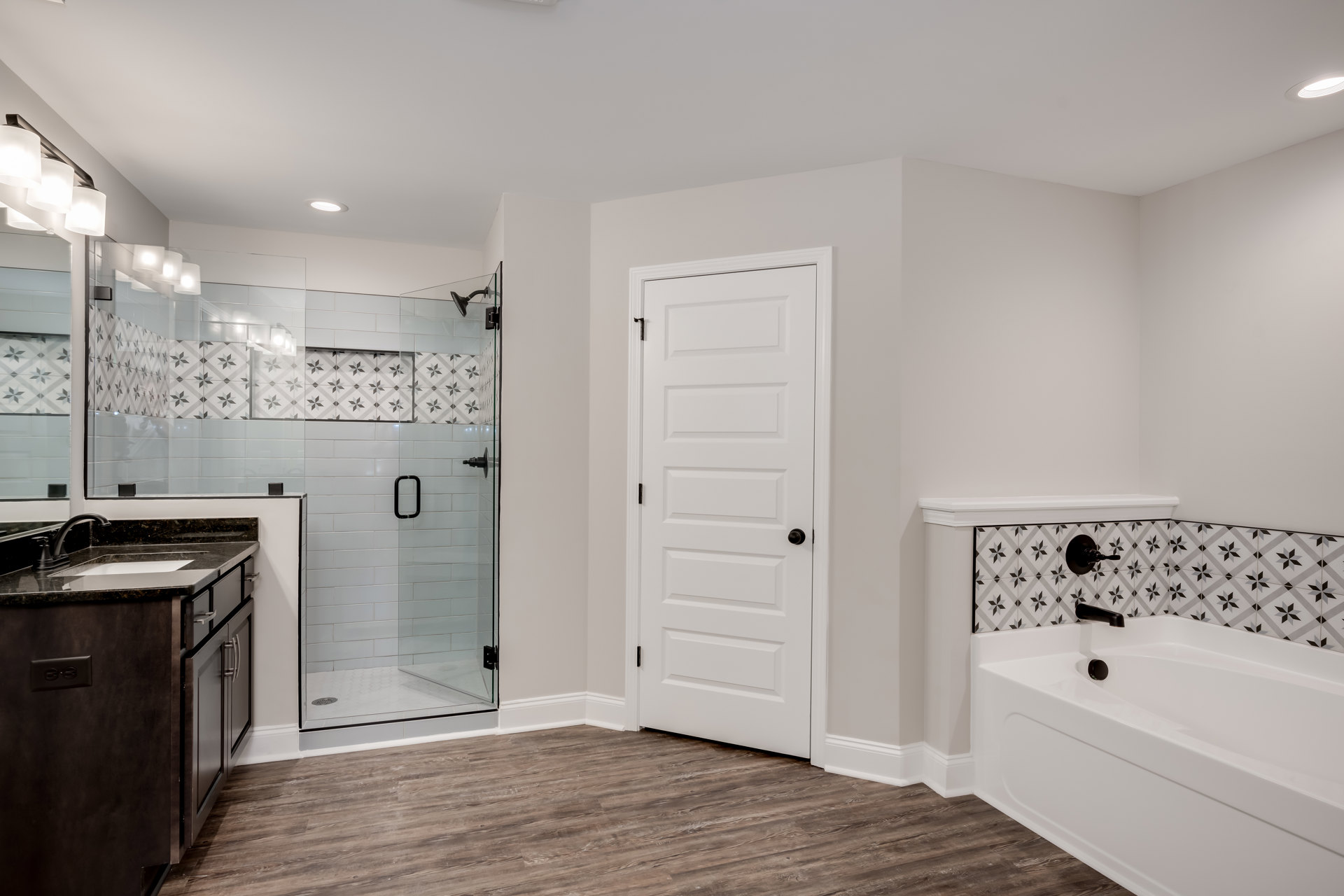 Bathroom featuring a glass shower door, white bathtub with black faucet, white door with black knob, tile wall with black hardware, and partial view of kitchen island with sink and