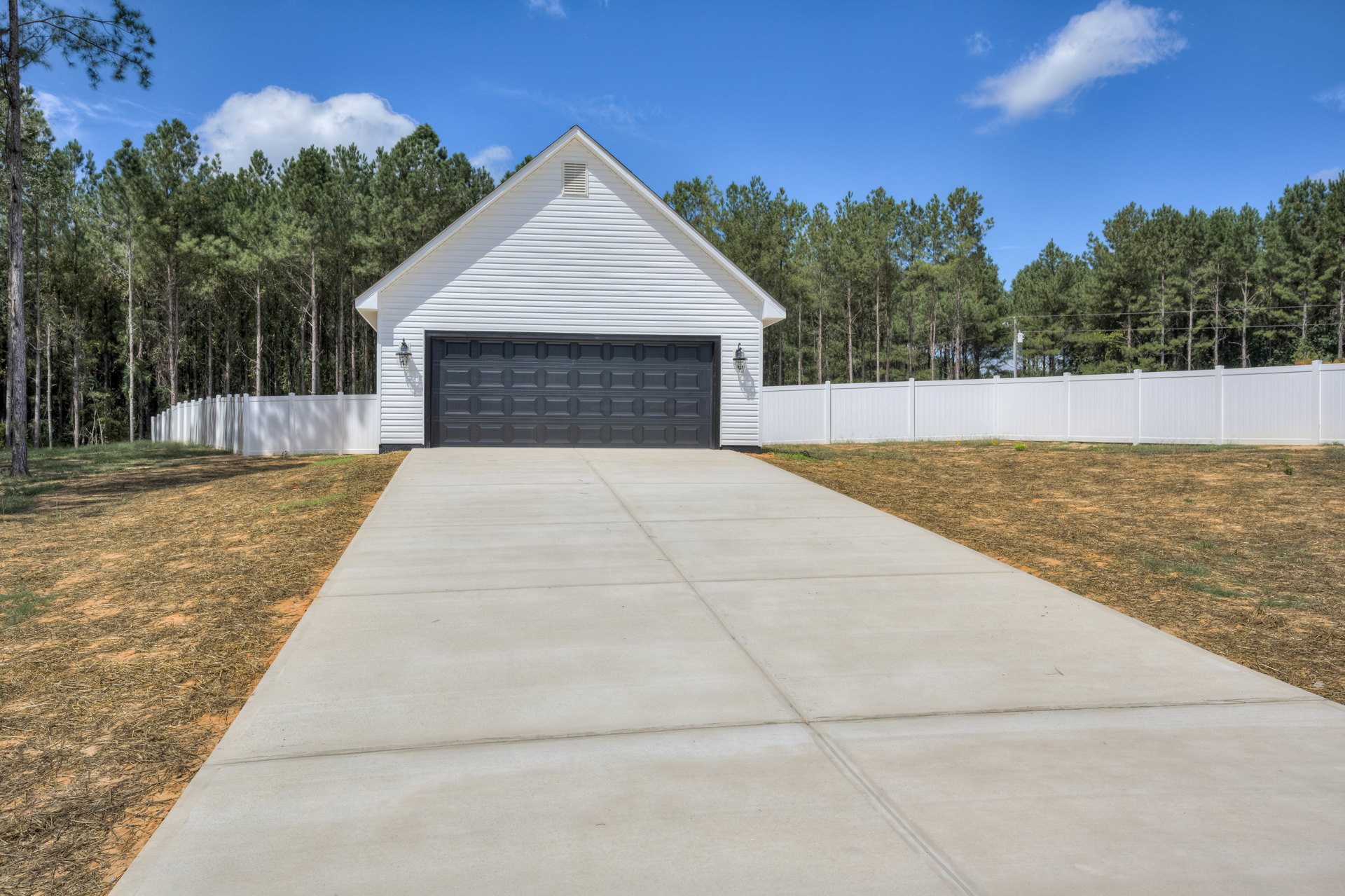 Concrete driveway bordered by grass leading to a white house with a black square-patterned garage door, white fence and mature trees in the background under a partly cloudy sky