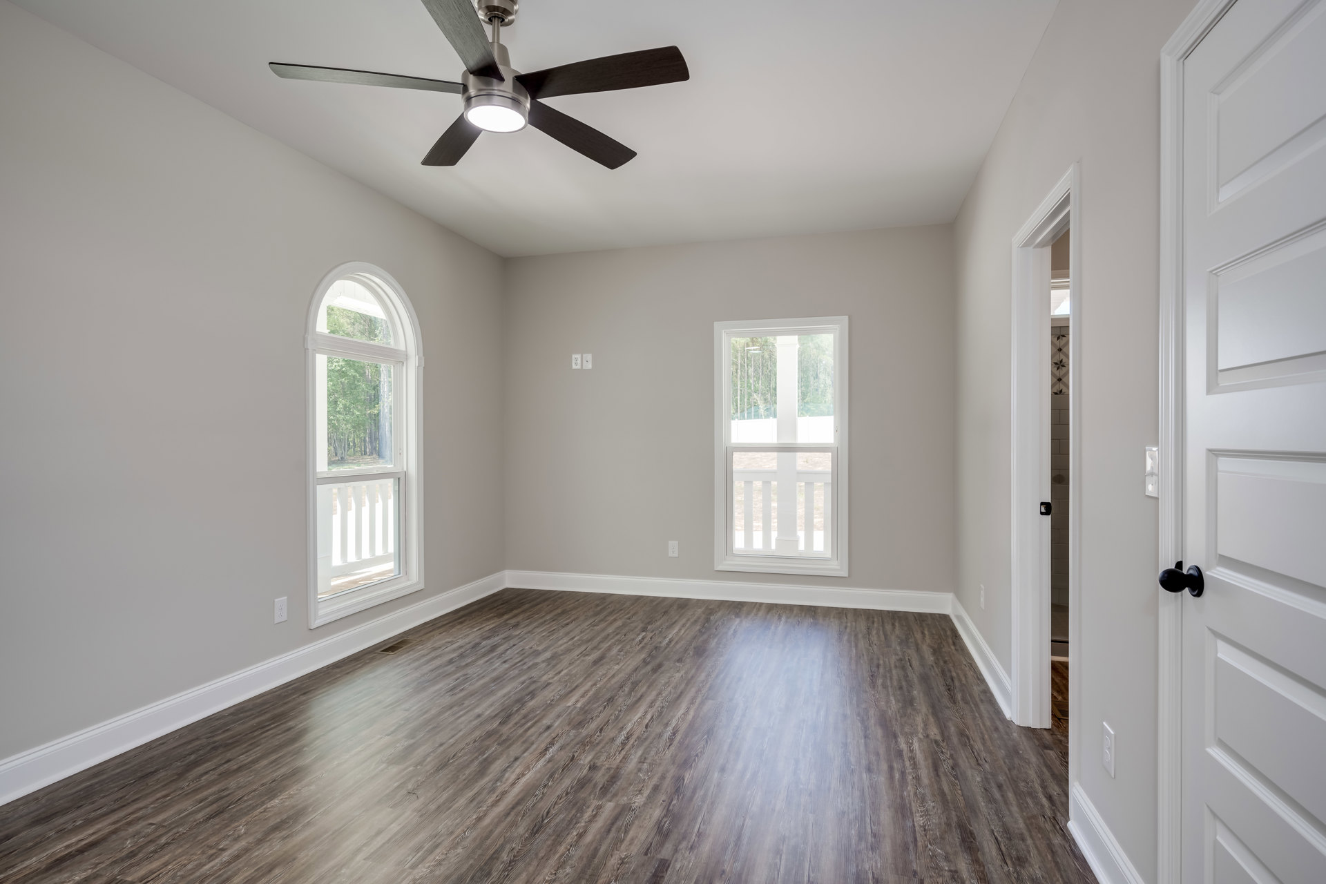 Ceiling fan with light fixture, white walls, wood flooring, large windows showing trees outside, white screen door with matching railing