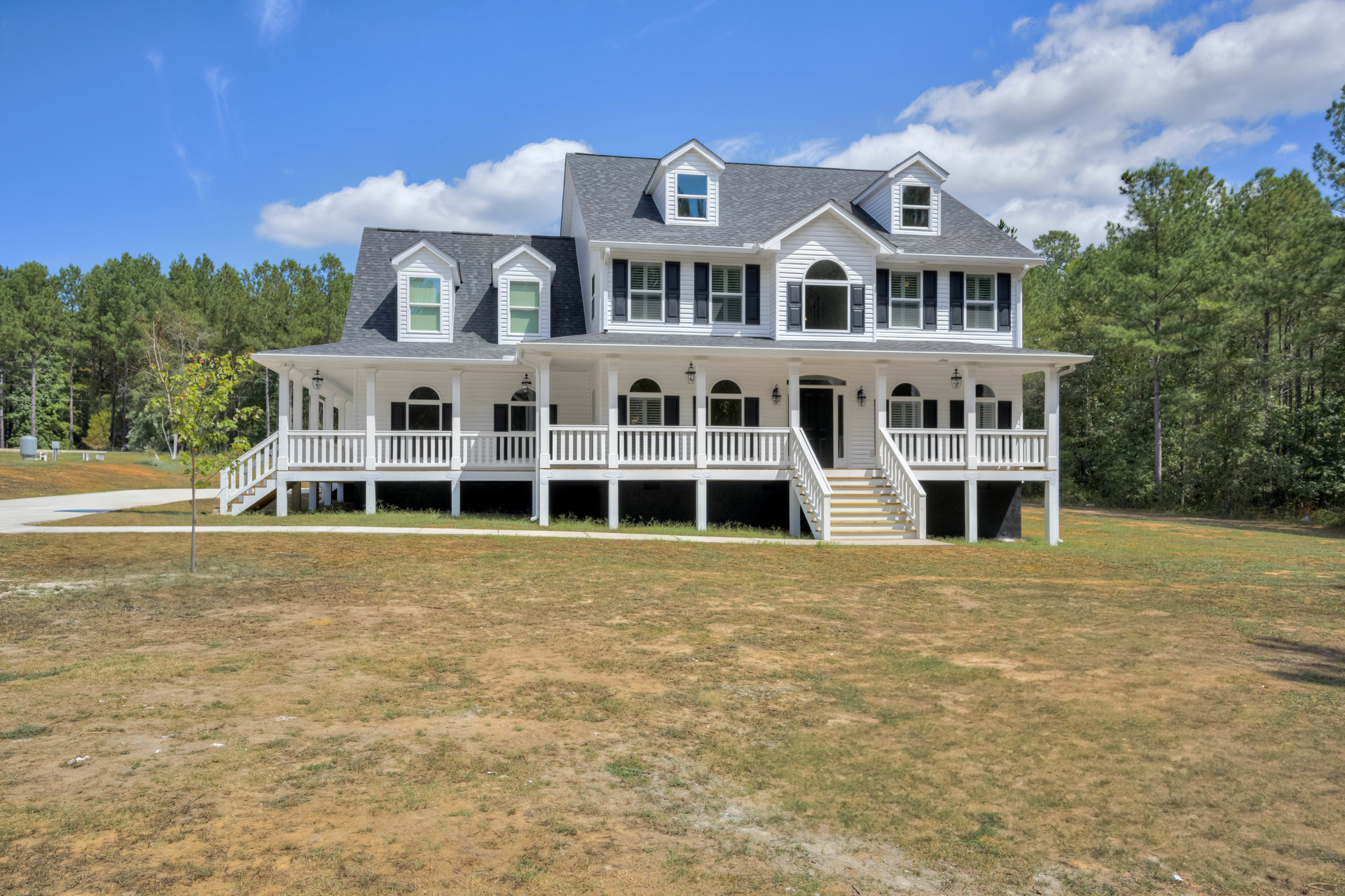 White two-story house with wide covered front porch, white railings, and steps leading to a grassy lawn; large windows reflect blue sky and clouds.