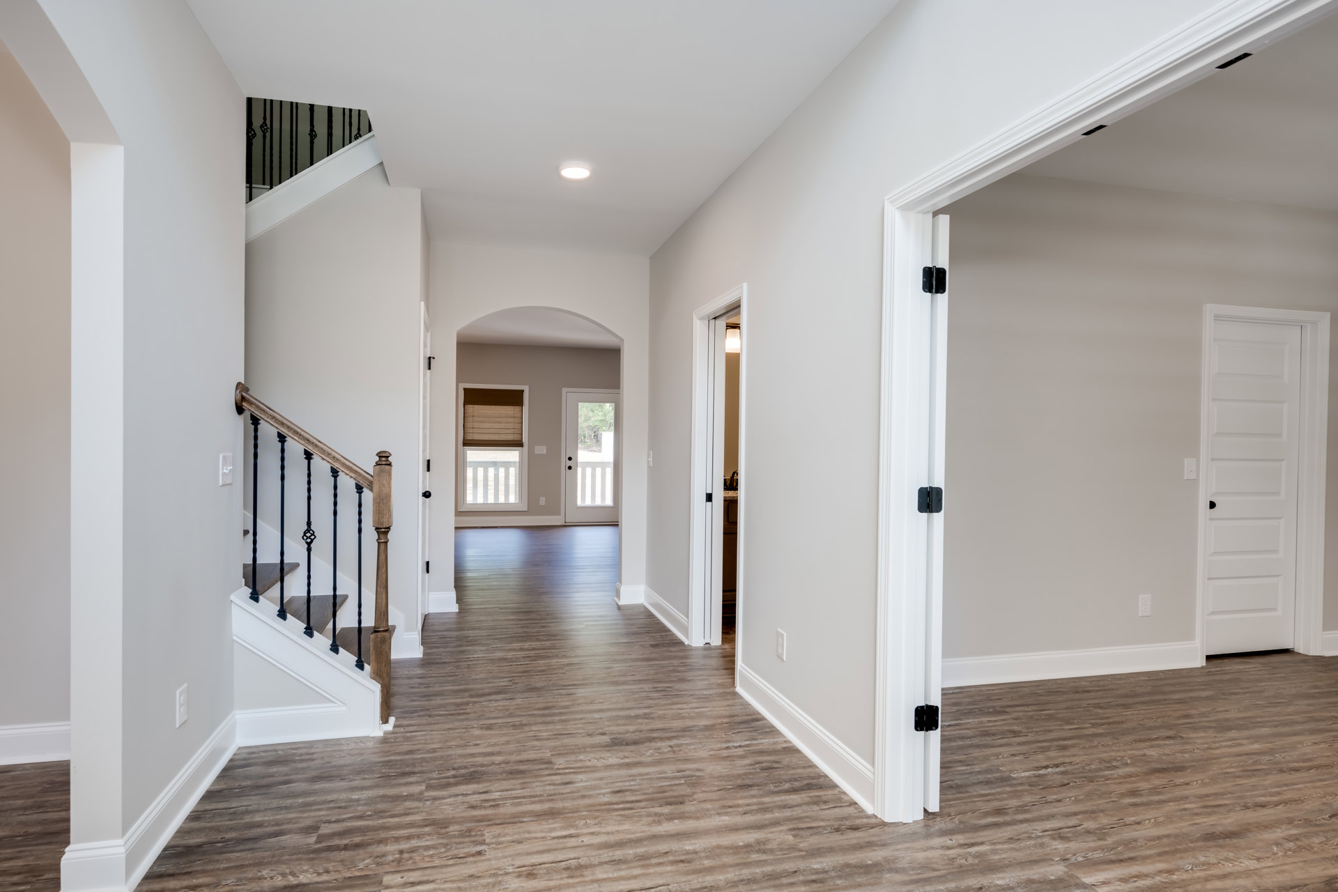 Hallway with light laminate flooring, white walls, wooden staircase with black railing, white door featuring black handle, window with brown shade, crown molding along ceiling