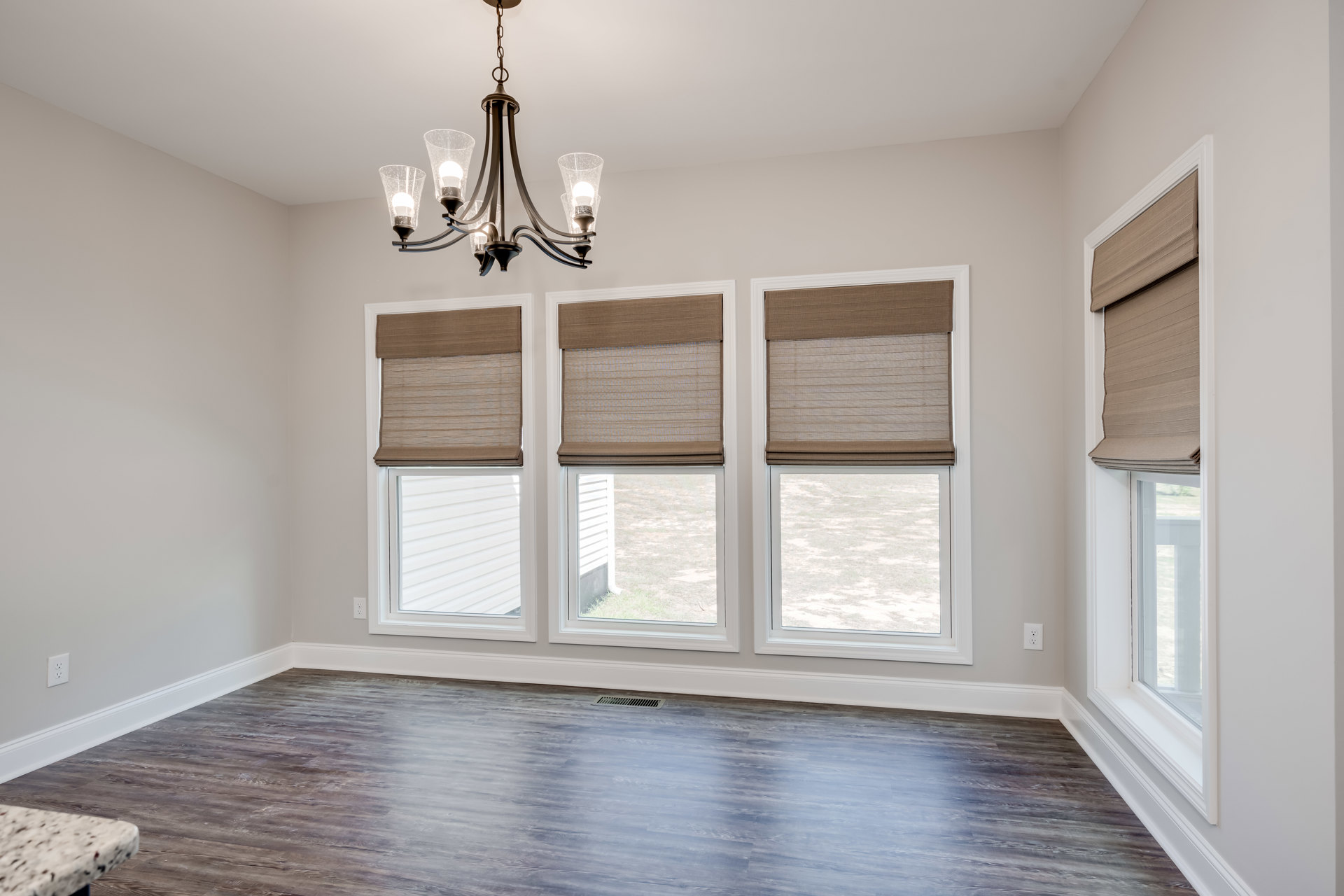 Neutral-toned room with large windows featuring brown blinds, white plaster walls, wood flooring with a floor vent, and a ceiling-mounted chandelier with clear glass shades