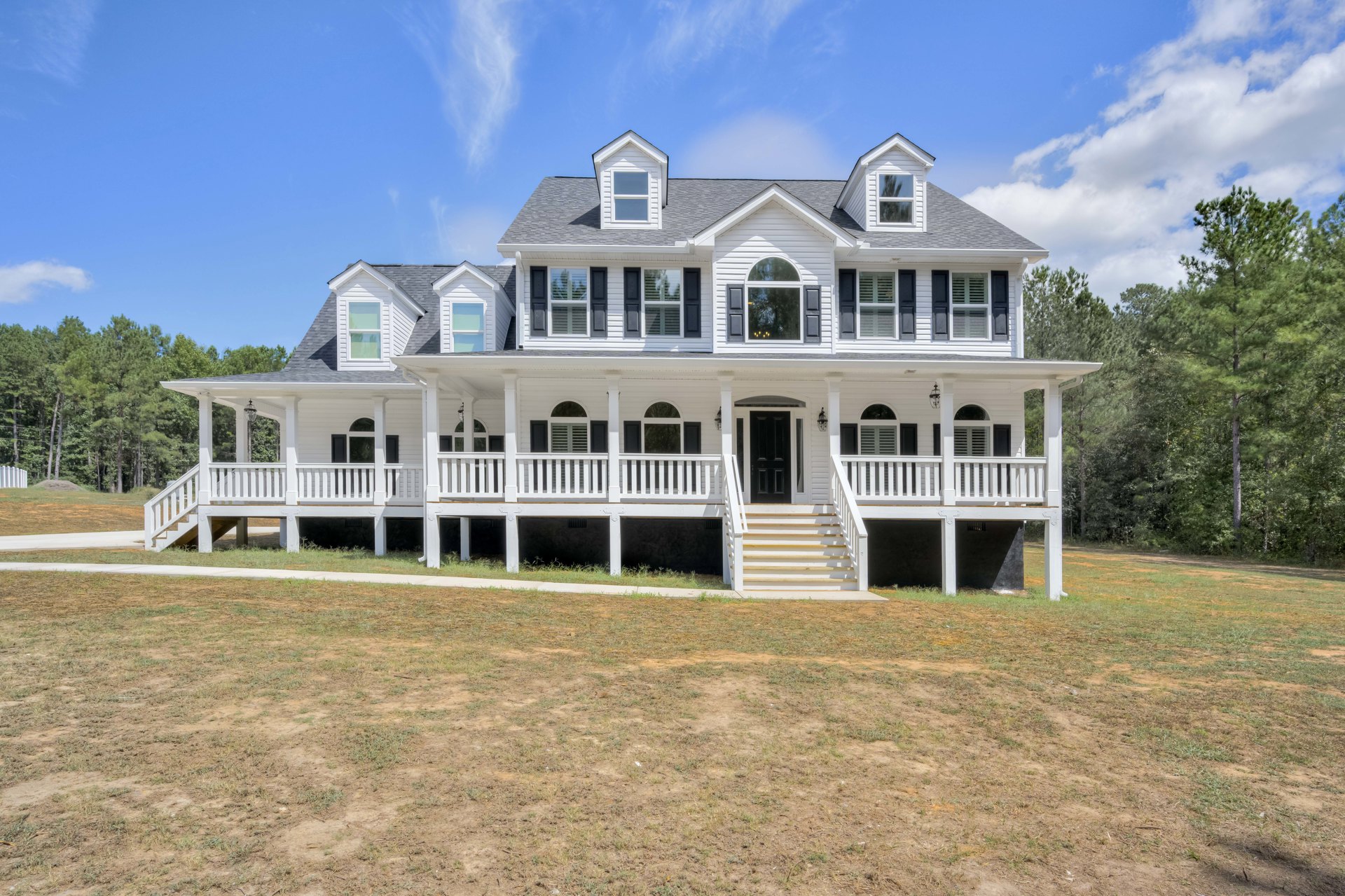 White two-story house with dormer windows, covered front porch, white railing, and expansive green lawn bordered by mature trees under a blue sky