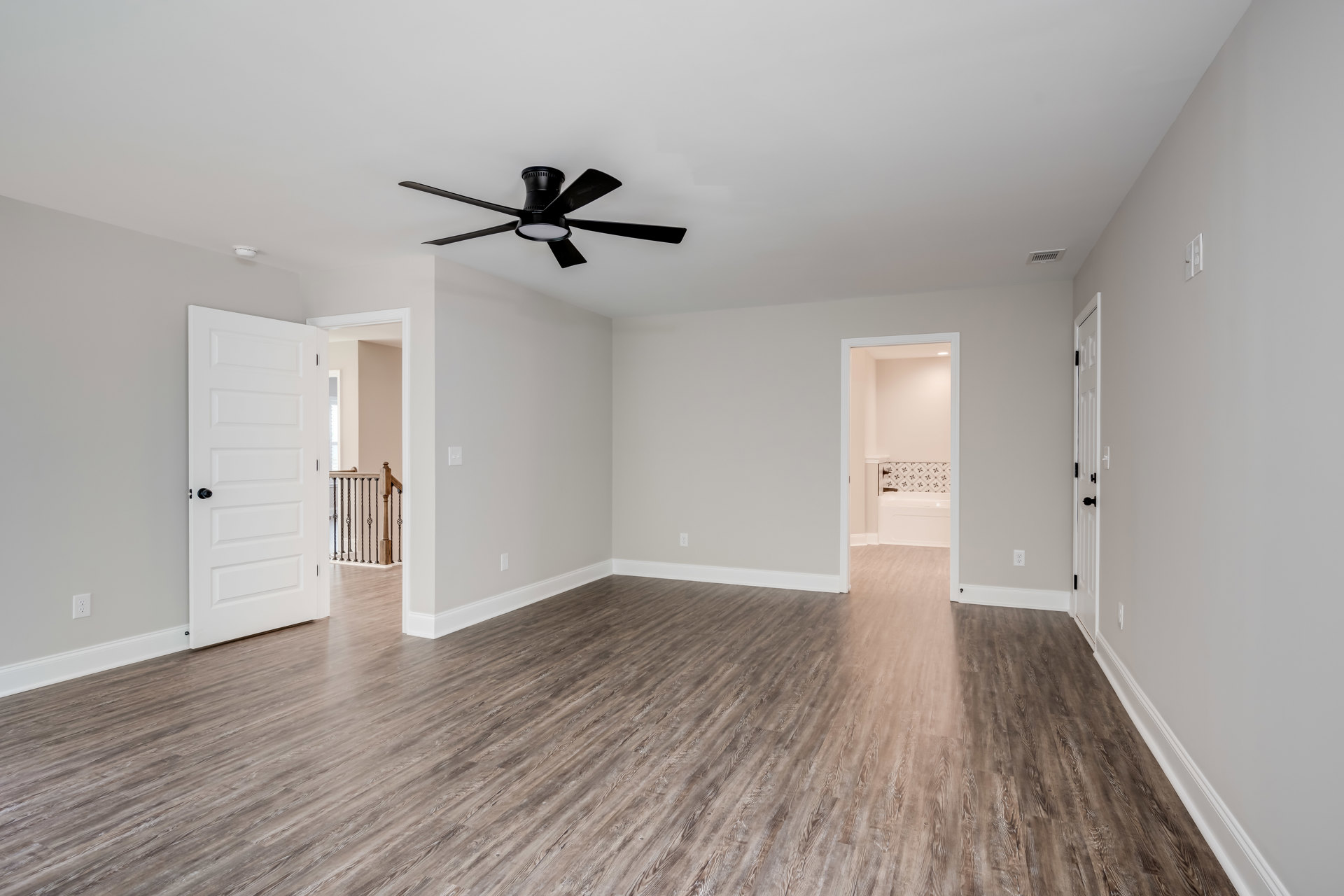 Bedroom with black ceiling fan, white walls, wood flooring, white door with black knob, and staircase with black metal balusters