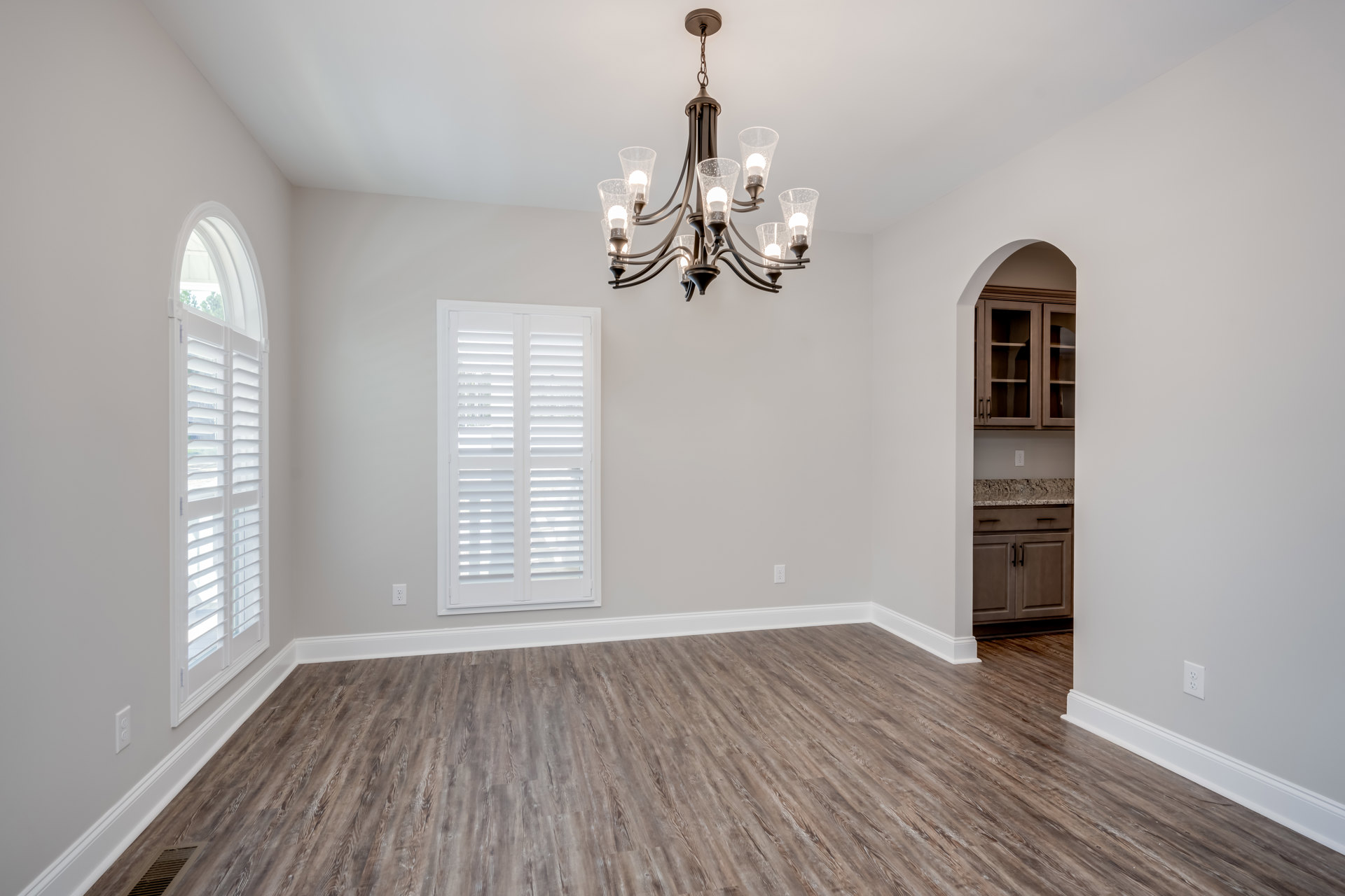 Chandelier with clear glass shades hanging from white ceiling, hardwood floor with white trim, large window with white blinds, light neutral walls