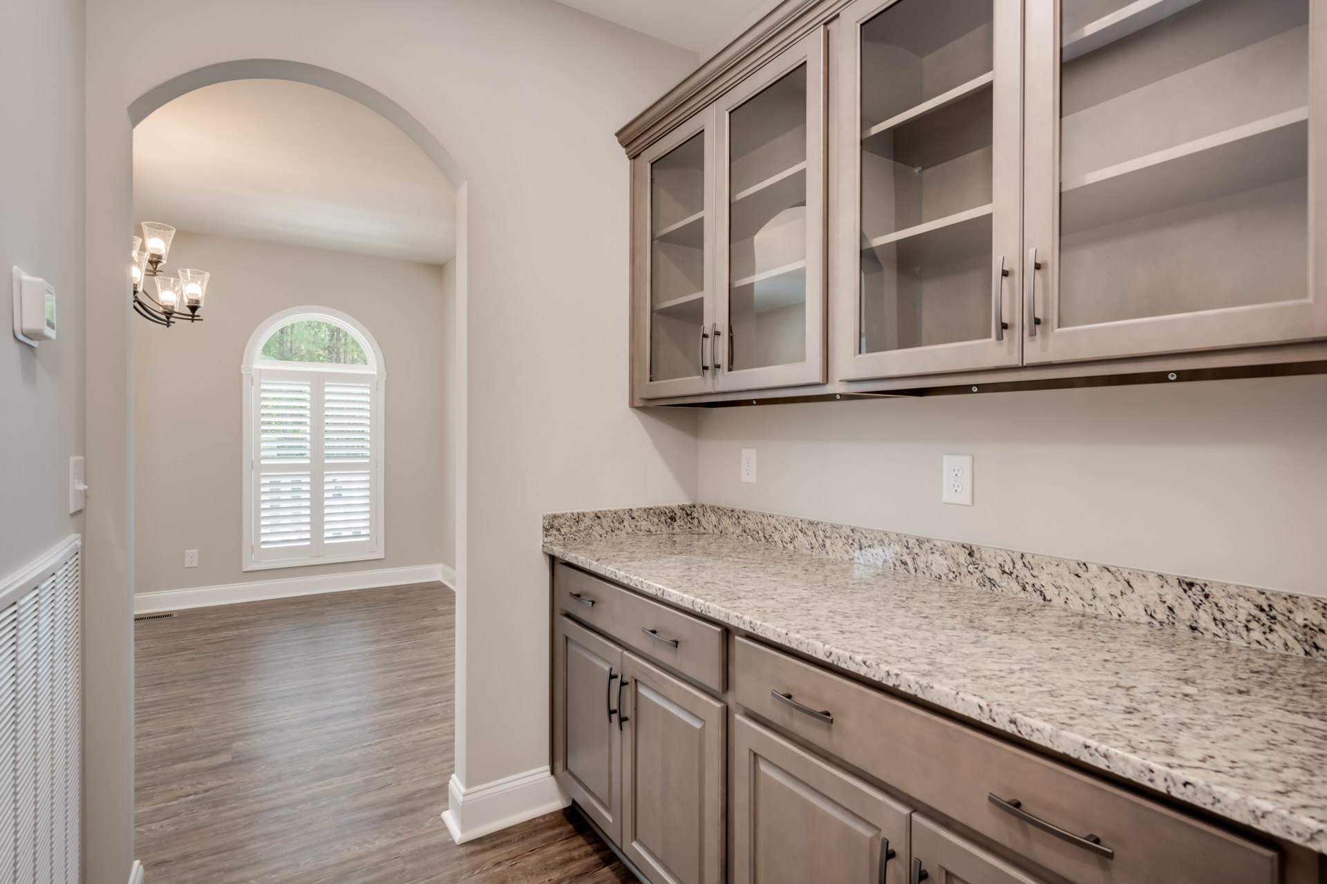 Marble countertops and wood cabinetry in a kitchen with glass doors, tile backsplash, stainless steel sink, and light fixture; wood flooring and white walls with vent visible.